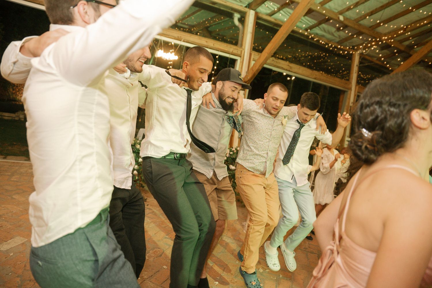 Wedding guests dance together on a rustic wooden floor under string lights at an outdoor reception venue.
