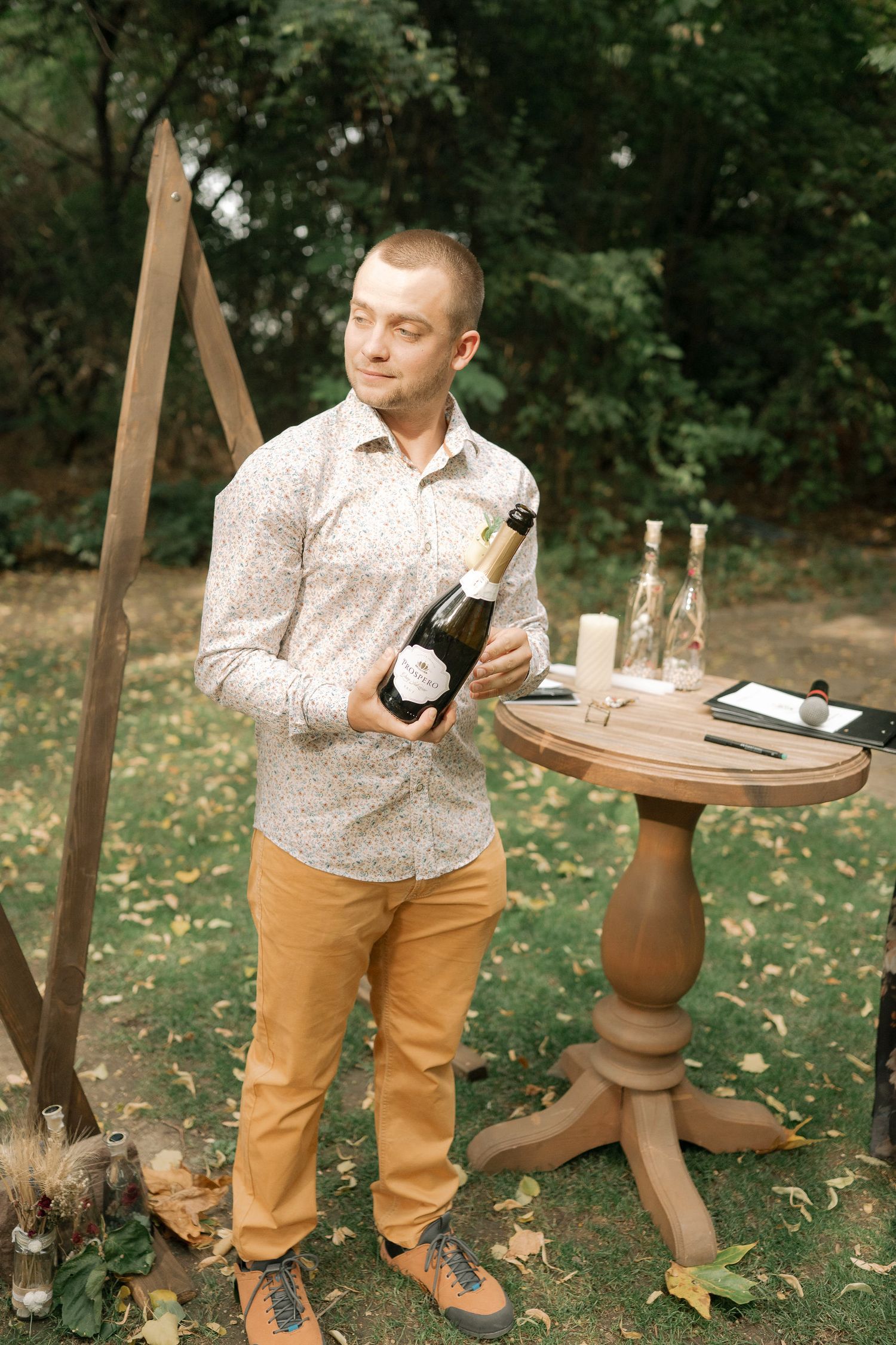 A person in a floral shirt and khaki pants holding wine bottles in an outdoor garden setting.