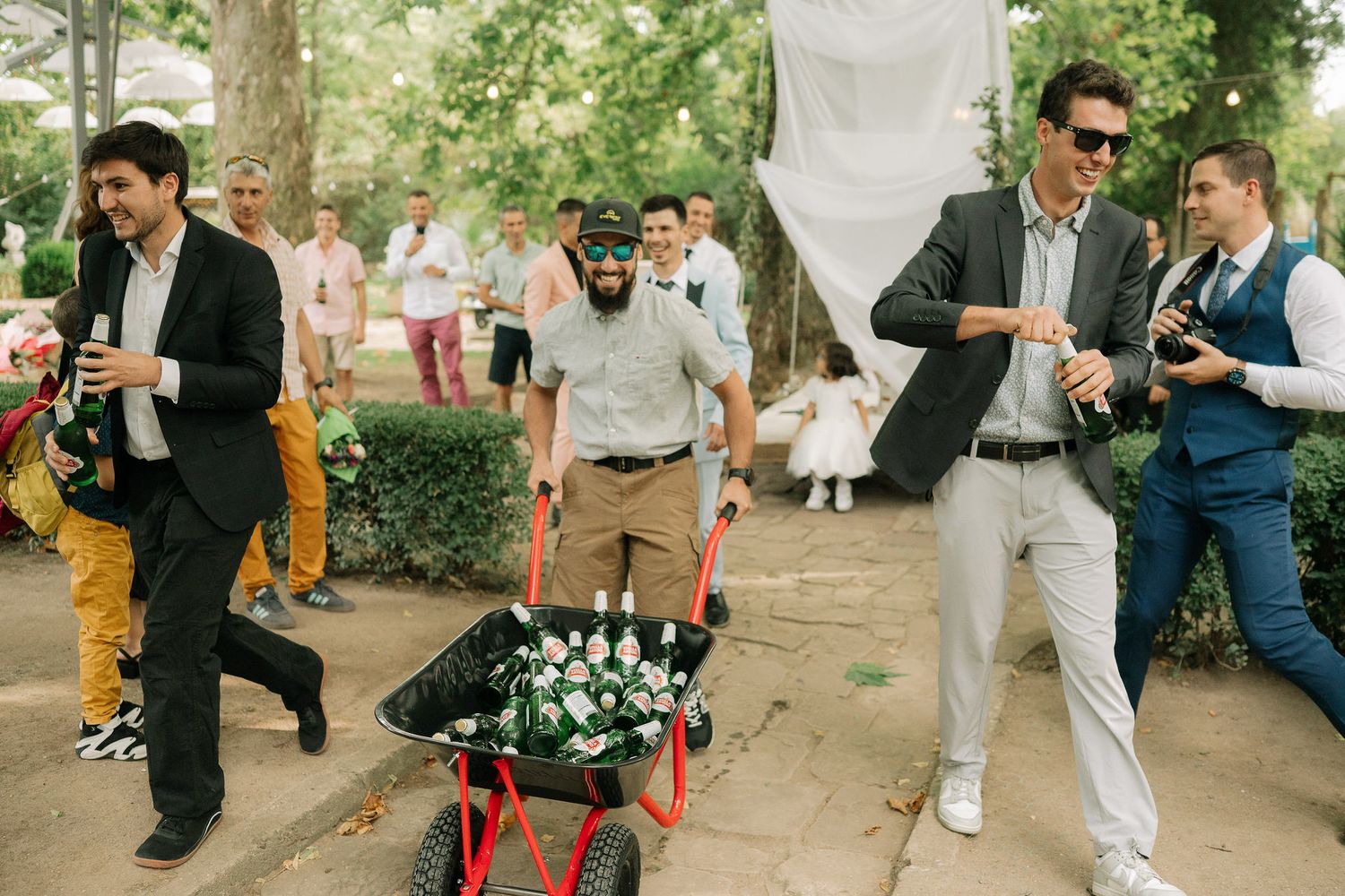 Guests in formal attire gather around red chairs in an outdoor garden setting during a celebration.