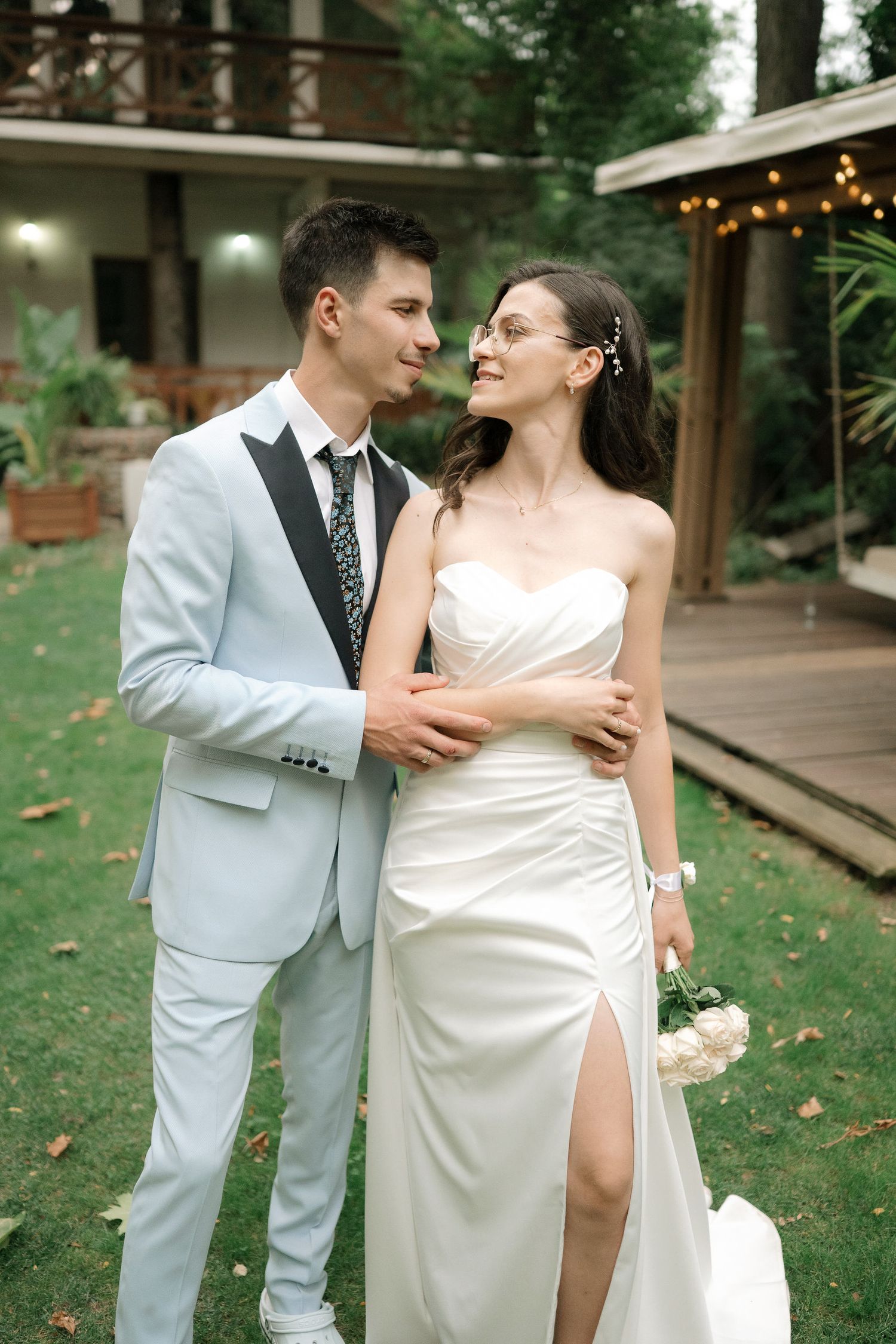 Newlywed couple shares romantic moments in an outdoor garden setting wearing white and grey formal attire.