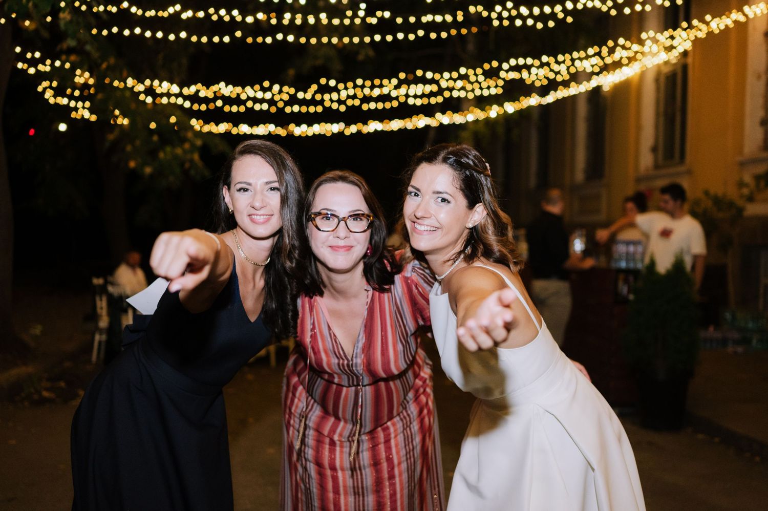 Three friends pose together at a nighttime outdoor party under twinkling string lights.