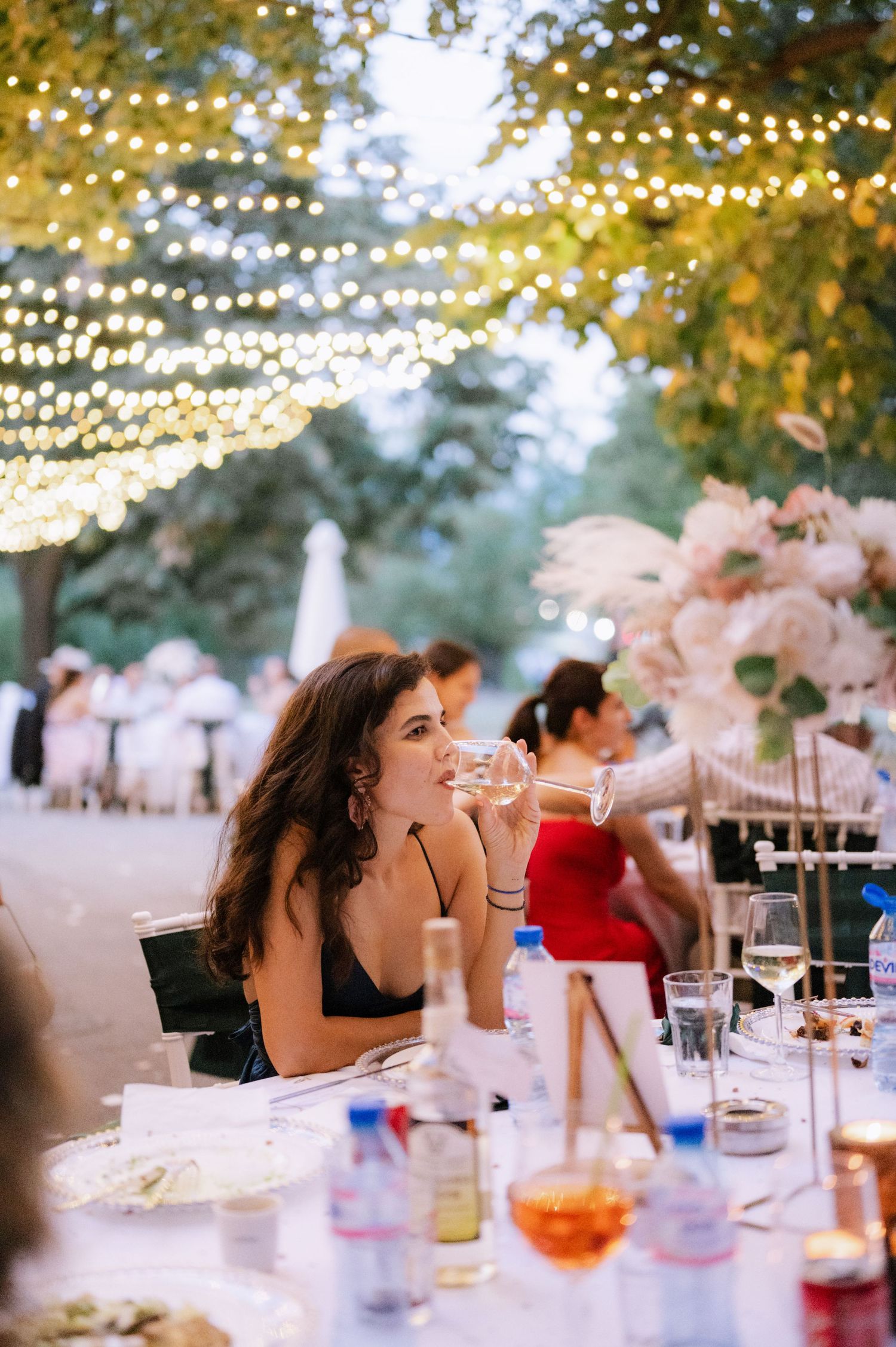 Guests enjoy an elegant outdoor wedding reception under twinkling string lights and floral decorations at dusk.