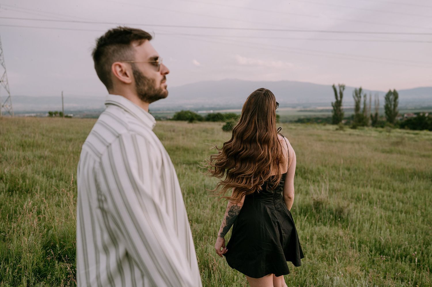 A couple in a grassy meadow shares a romantic moment against a scenic mountain landscape at sunset.