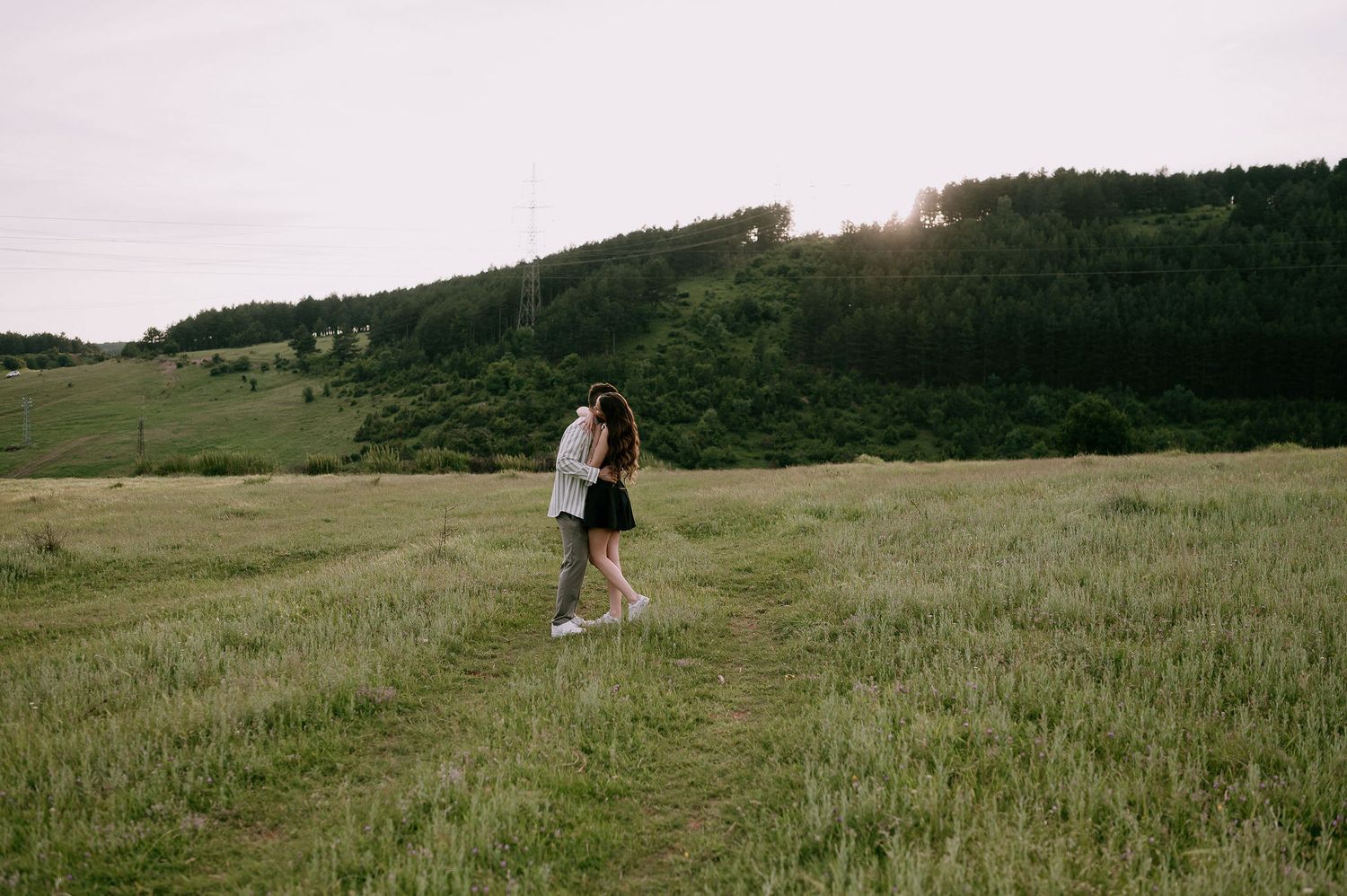 A couple embraces in a serene meadow with rolling hills and forest in the background at sunset.
