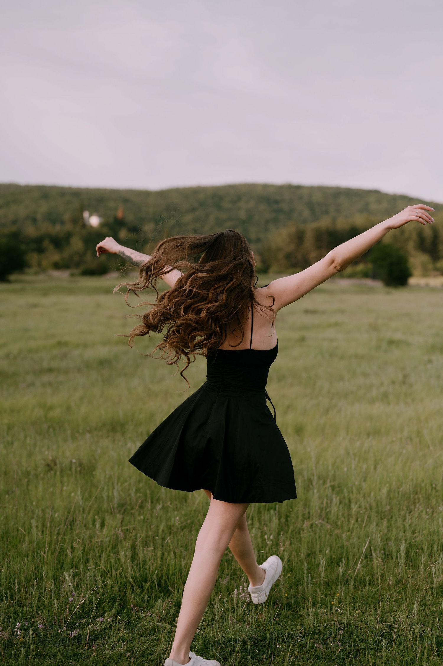 A person in a black dress twirls joyfully in a green meadow with mountains in the background.