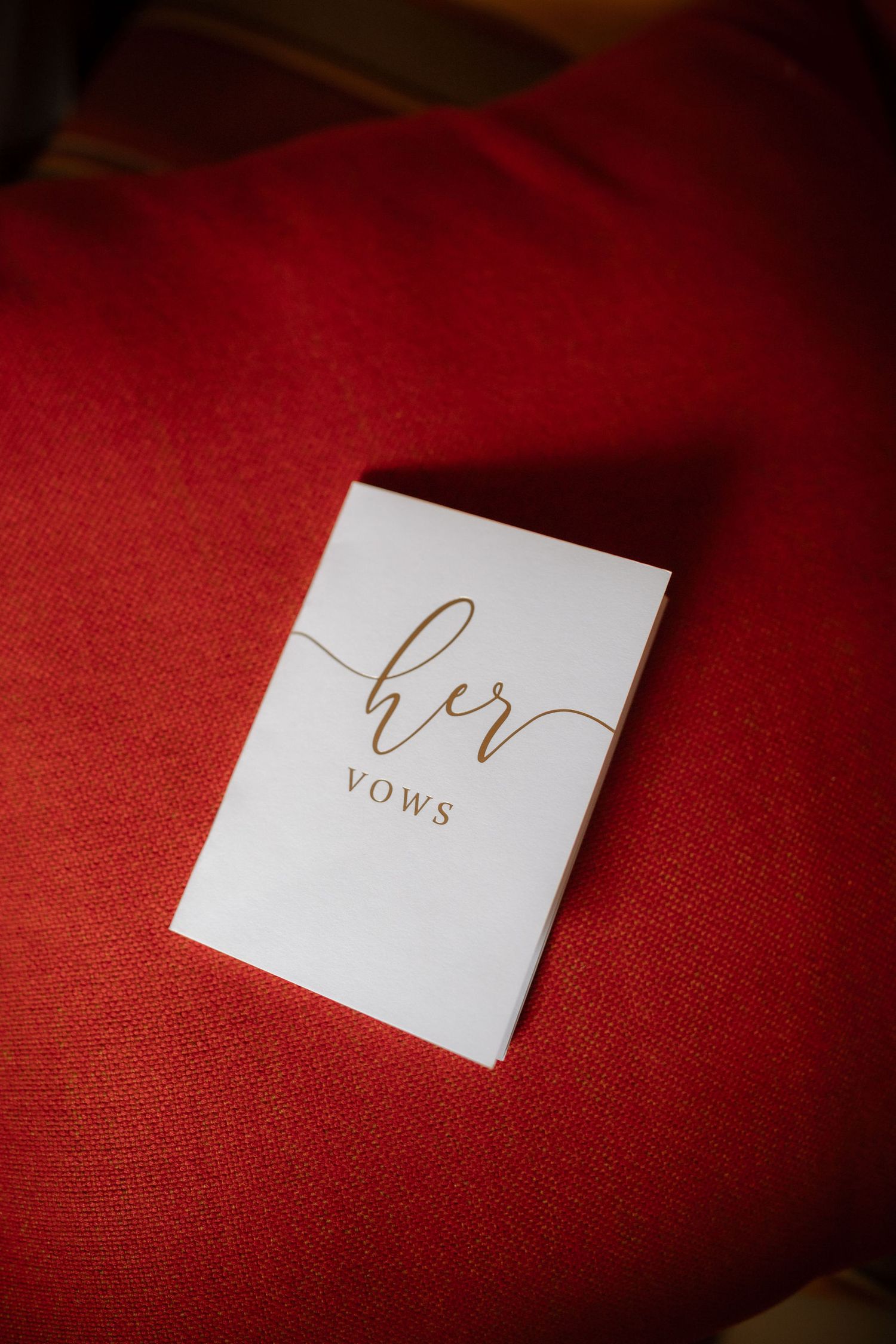 Wedding vows card with elegant gold calligraphy text on white paper against a deep red textured background.