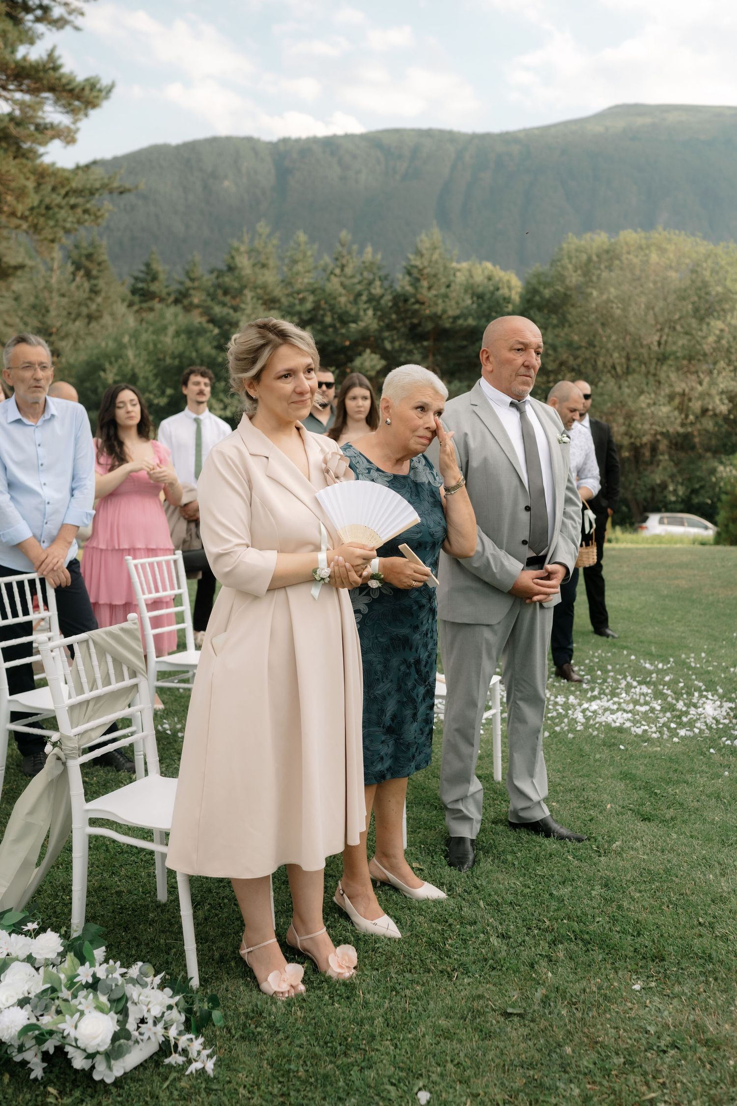 Wedding guests in formal attire sit on white chairs on a grassy lawn with mountains visible in the background.