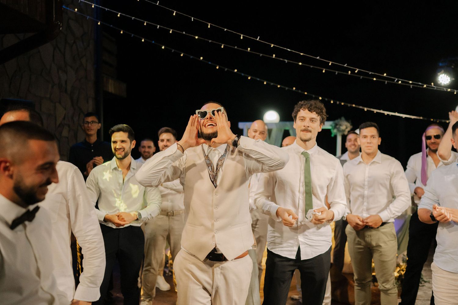 Wedding party members in light-colored suits dance together in a line during a reception celebration.