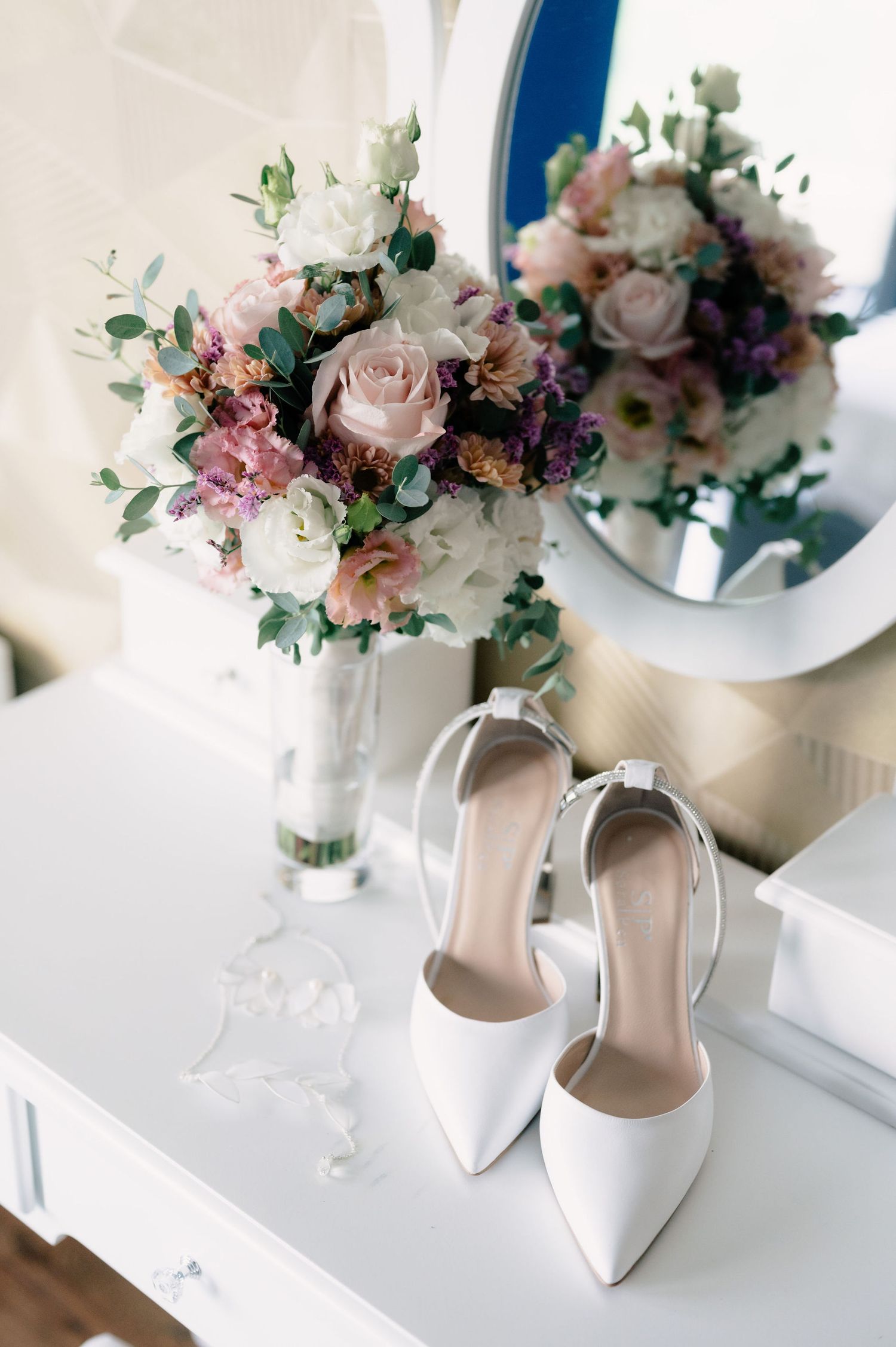 Elegant white bridal heels next to a romantic bouquet of blush roses and purple flowers in a vase.