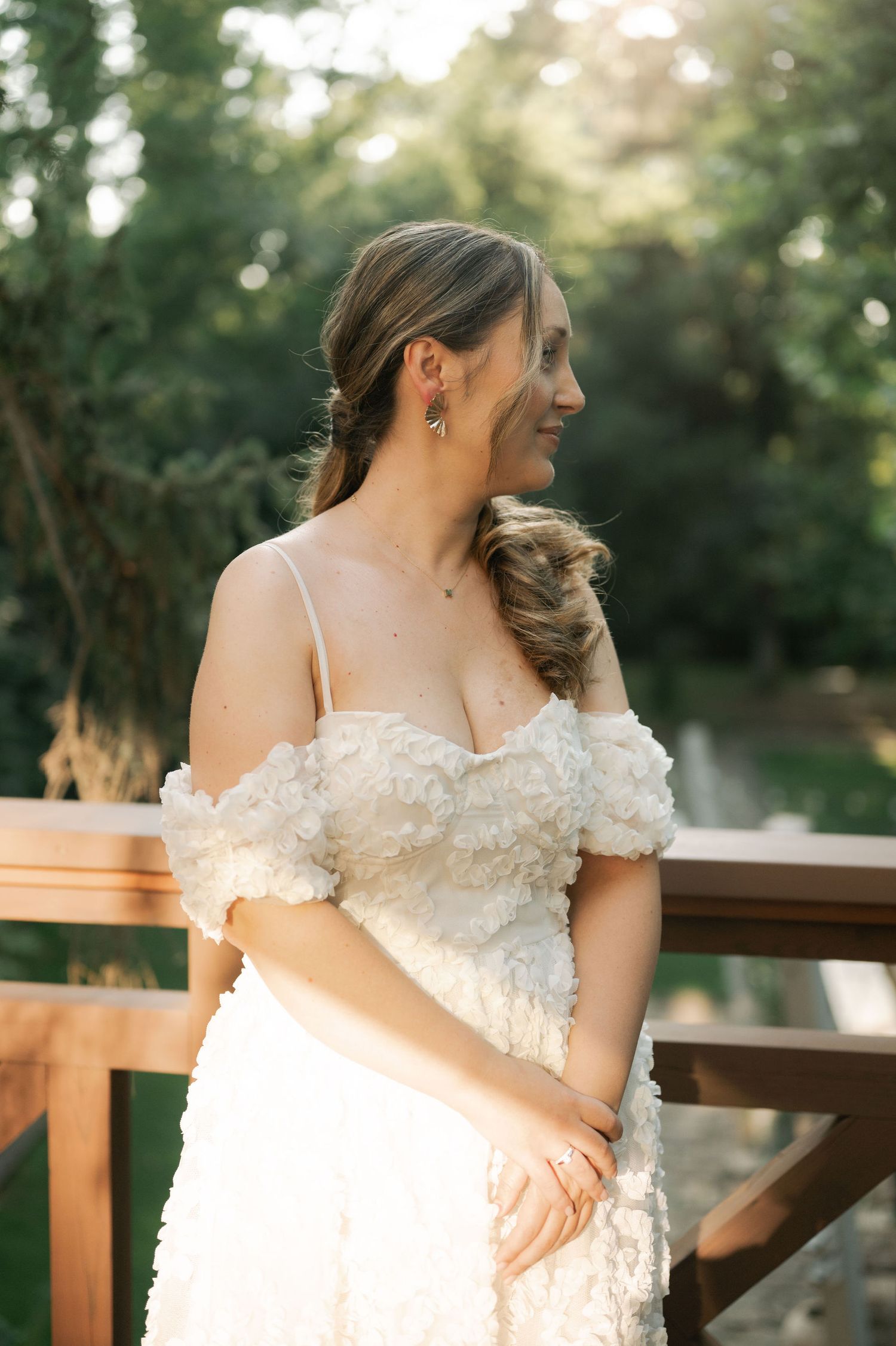 A bride in a white lace gown poses outdoors against a wooden railing during golden hour.