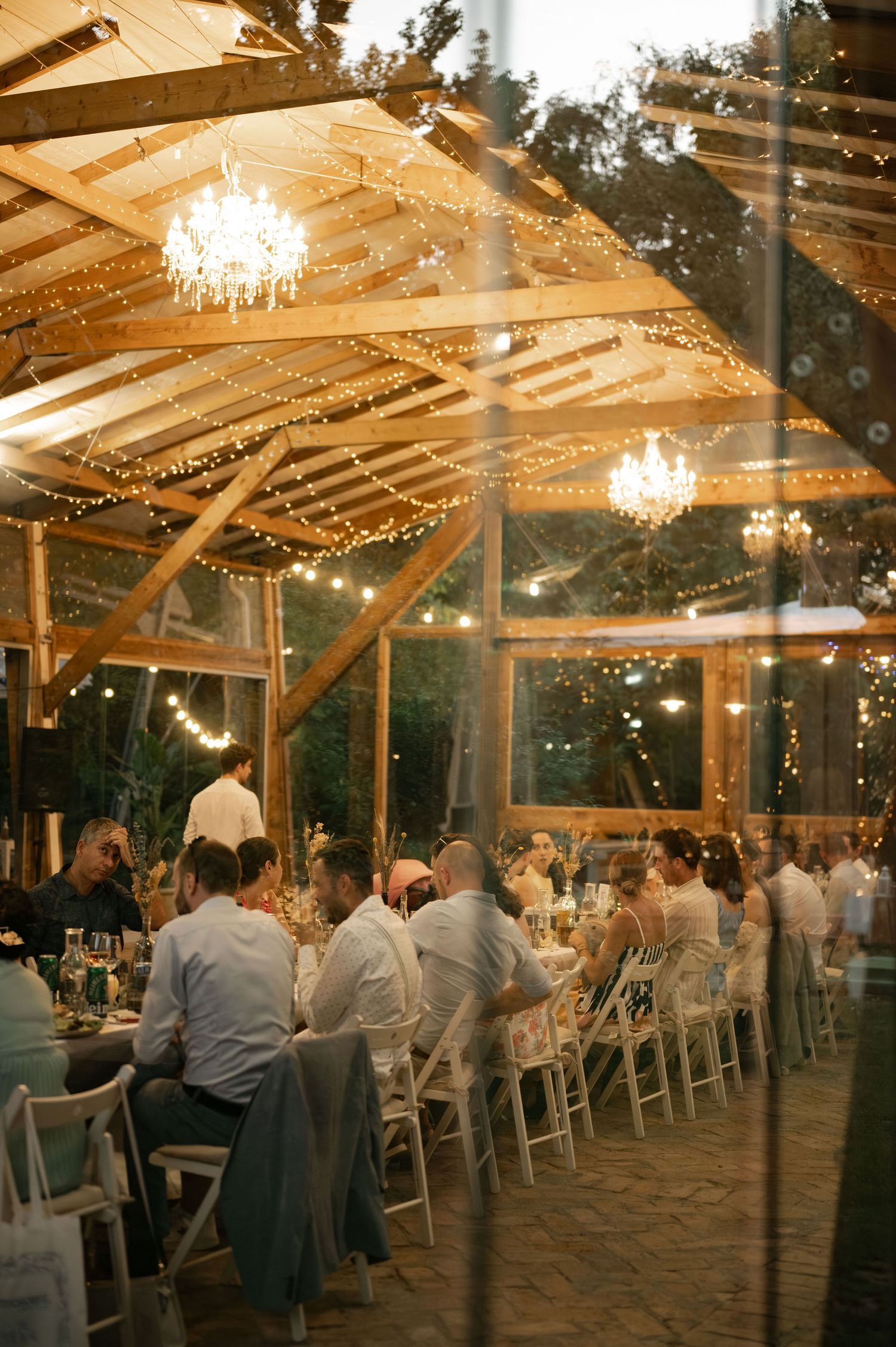 Elegant wedding reception in a glass-walled pavilion with twinkling lights and chandeliers creating a romantic atmosphere.