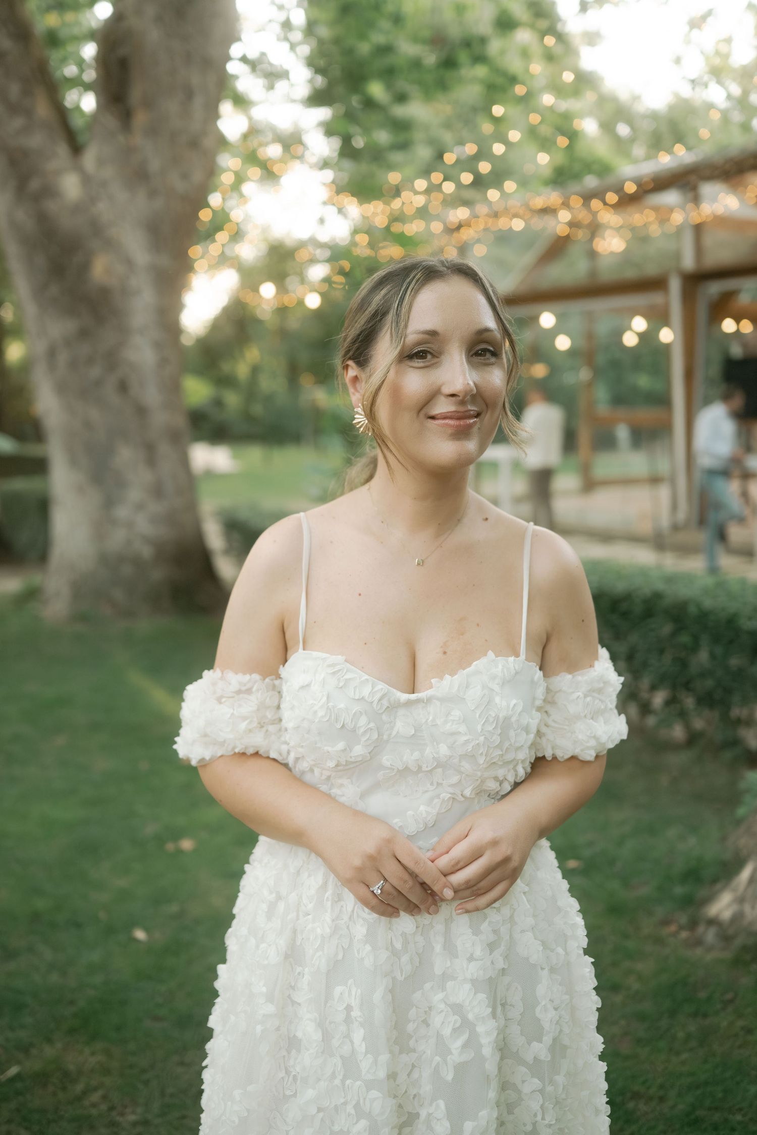 A bride in a white off-shoulder wedding dress stands in a garden setting with string lights and trees in the background.