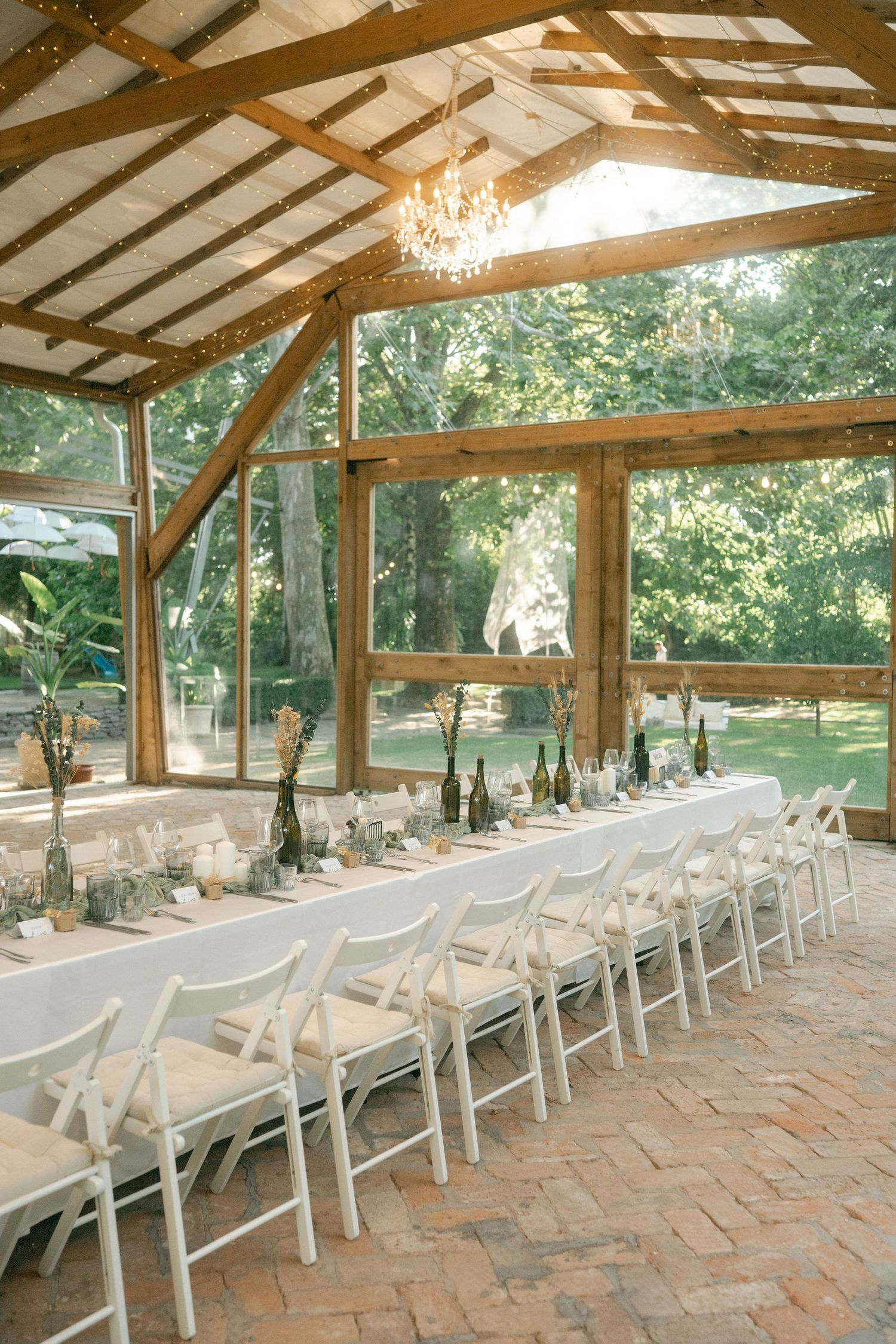 Rustic outdoor covered pavilion with rows of white chairs set up for an event on a brick patio surrounded by trees.
