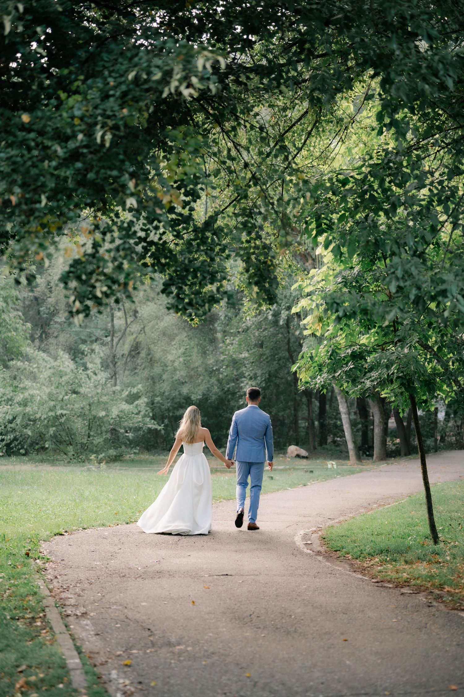 A couple in wedding attire walks along a winding dirt path through a lush green park on their special day.