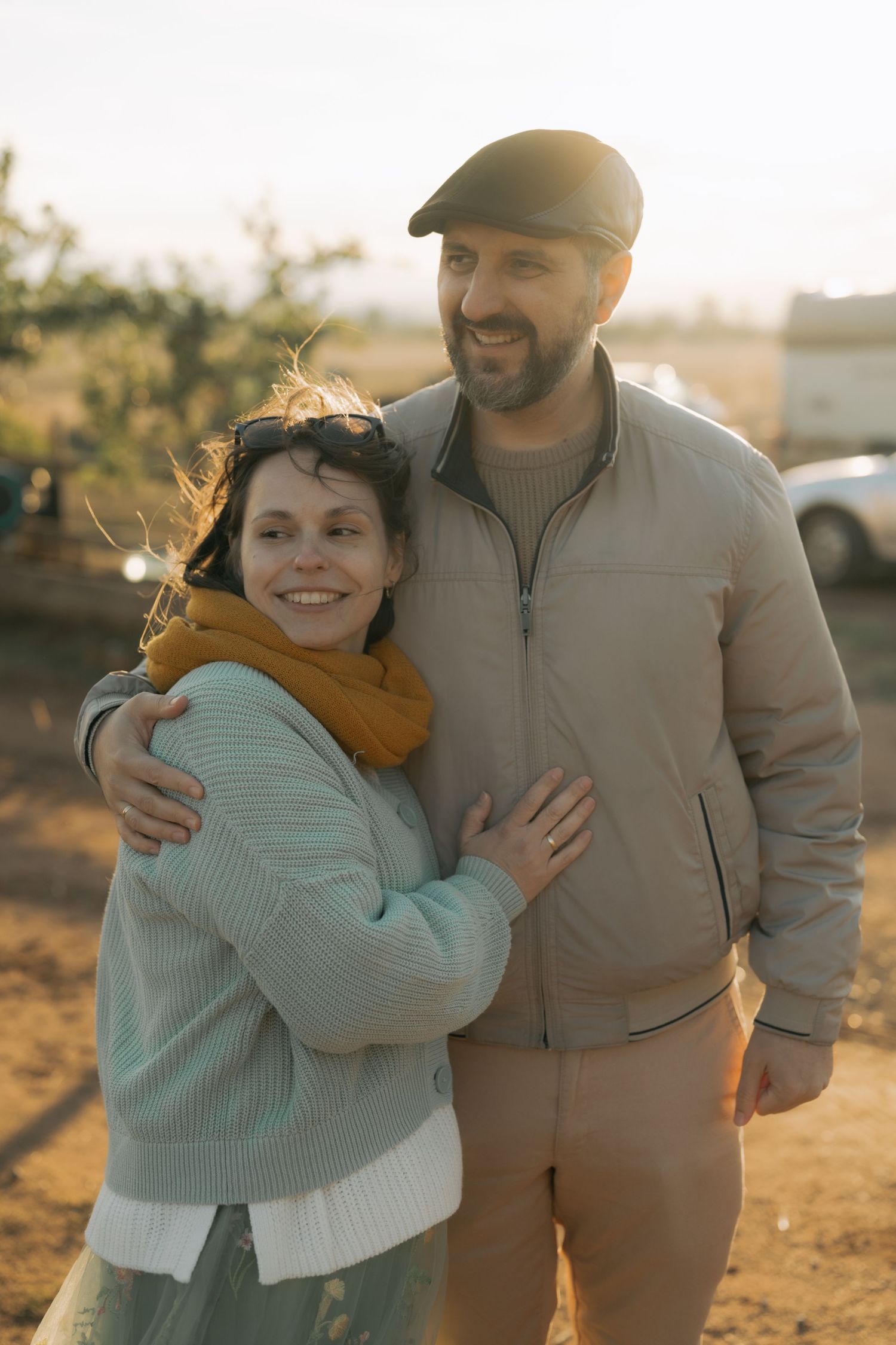 Couple embraces outdoors at sunset wearing casual fall attire including a flat cap and sweaters.