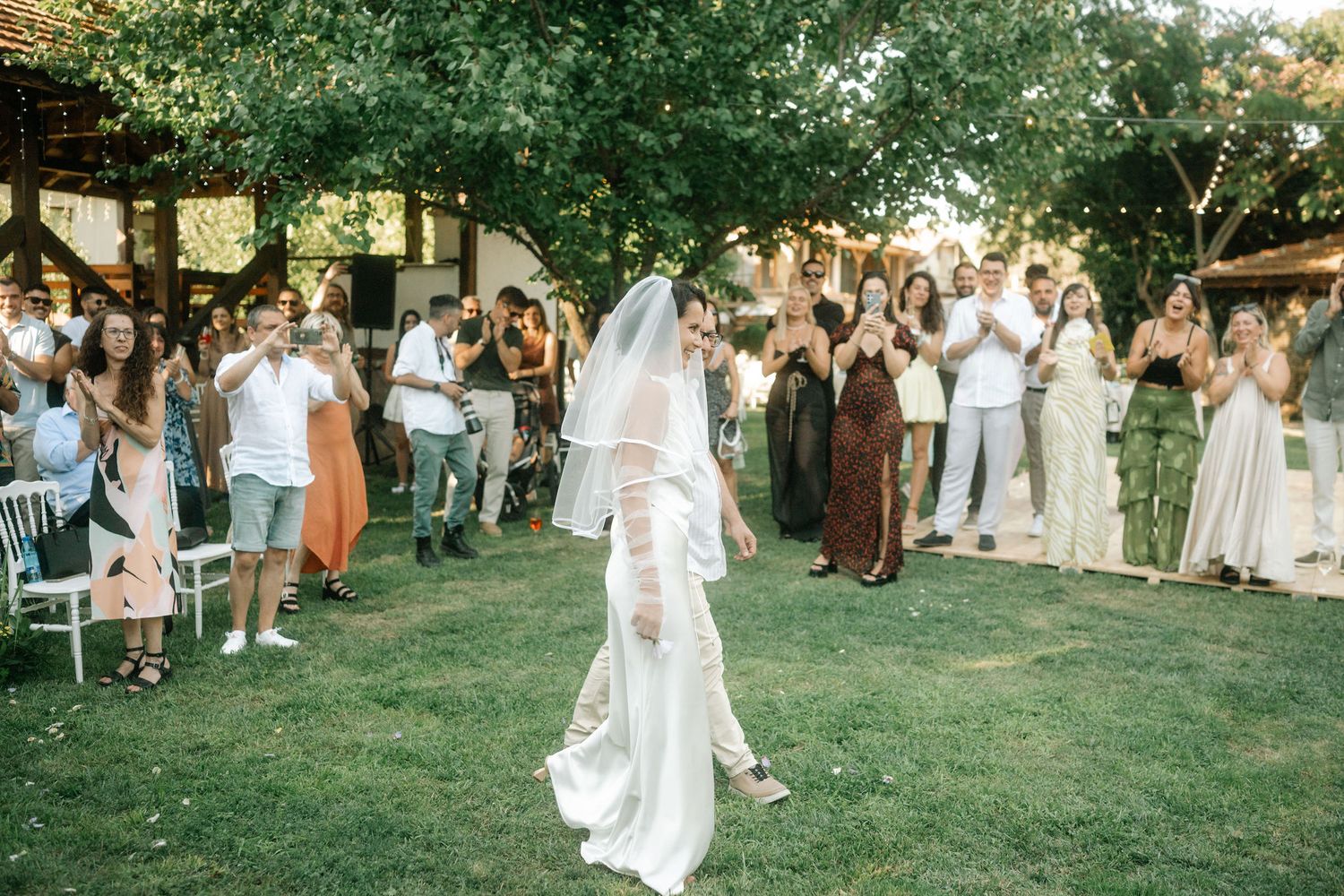 Wedding celebration on a green lawn with guests gathered around white chairs under trees.
