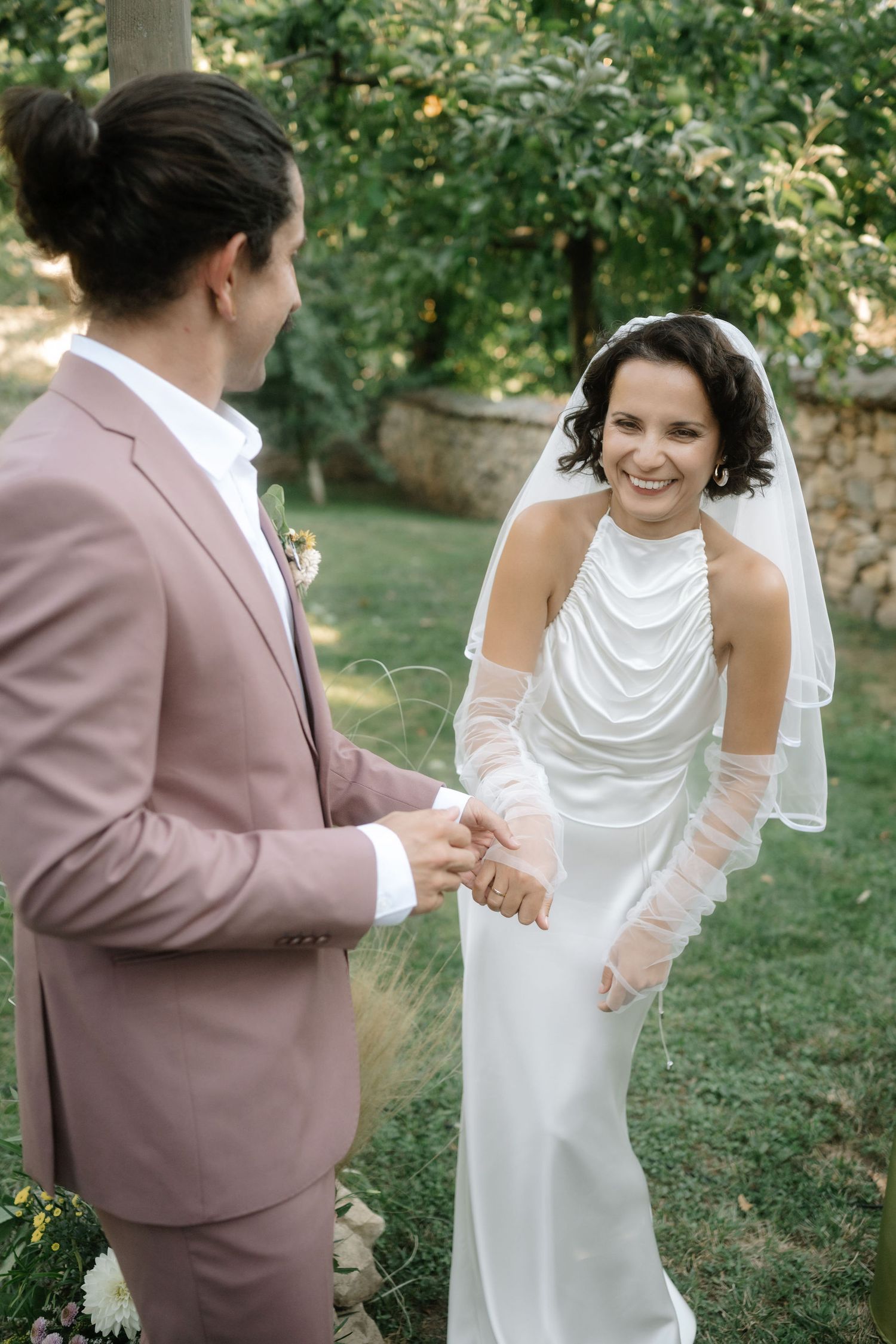 A couple shares a joyful moment during their outdoor garden wedding ceremony, with the bride in white and groom in dusty pink.