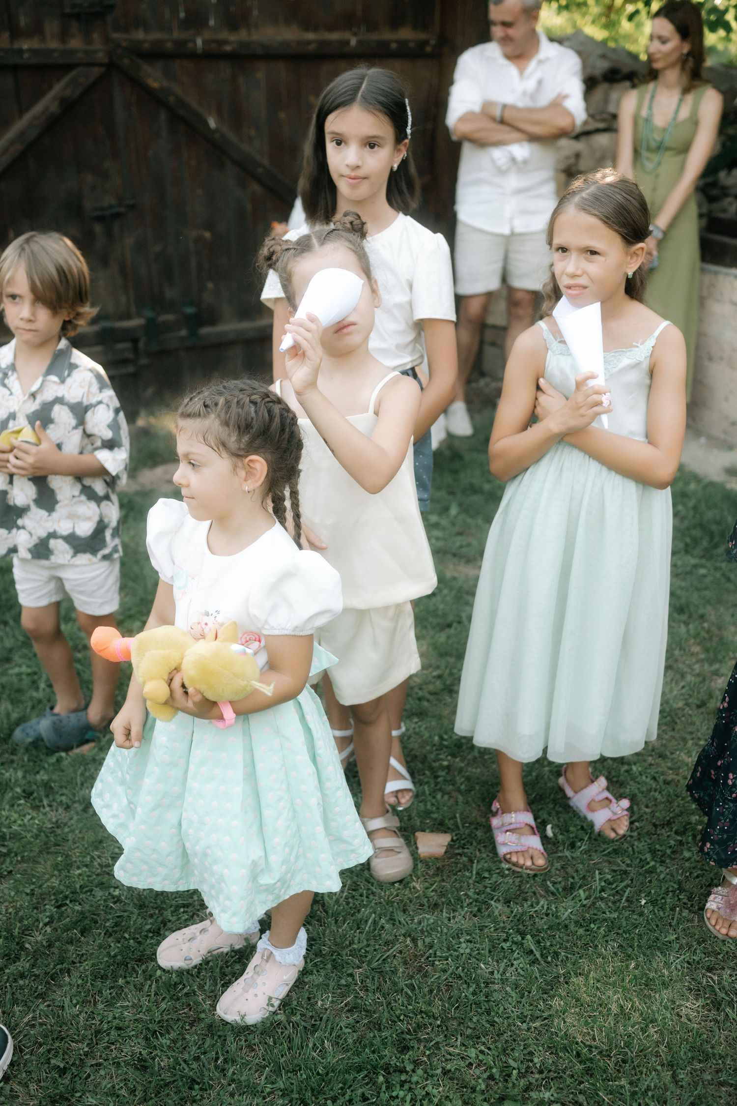 Children in white tops and mint green skirts gather on a grassy lawn during an outdoor event.