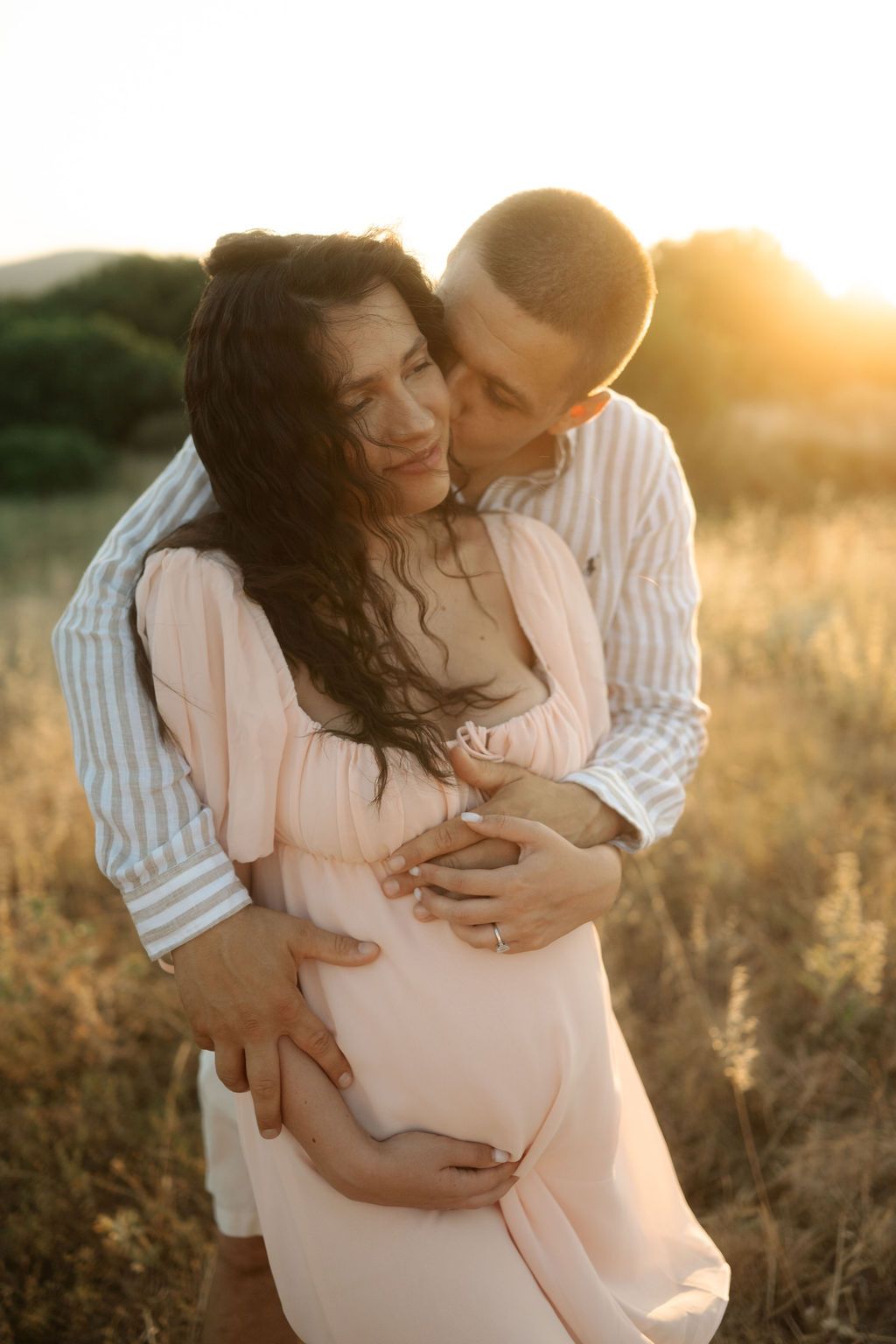 Romantic couple shares intimate moment in field during warm sunset while wearing white and striped clothing.