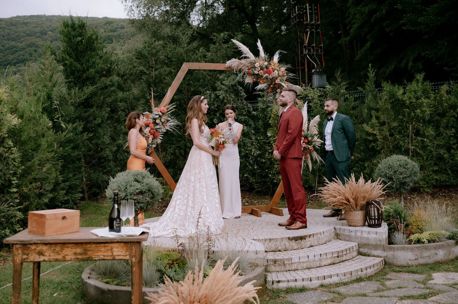Outdoor mountain wedding ceremony with rustic decor featuring pampas grass and wooden arch against evergreen forest backdrop.