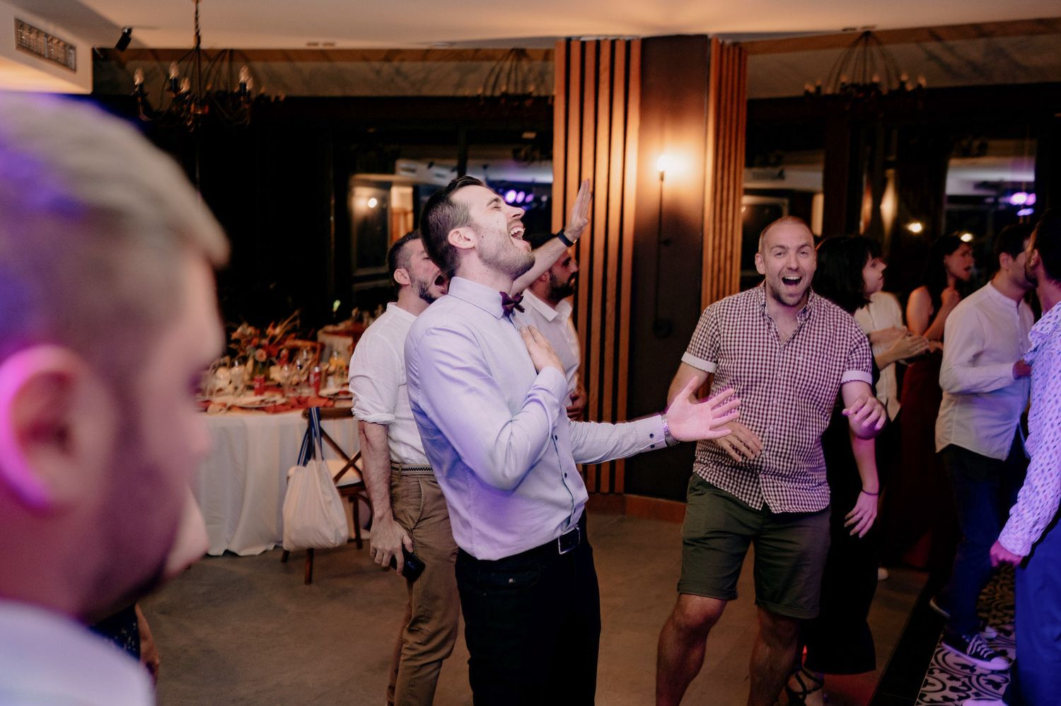 People dancing and enjoying themselves at an indoor wedding reception with tables and decorations in the background.
