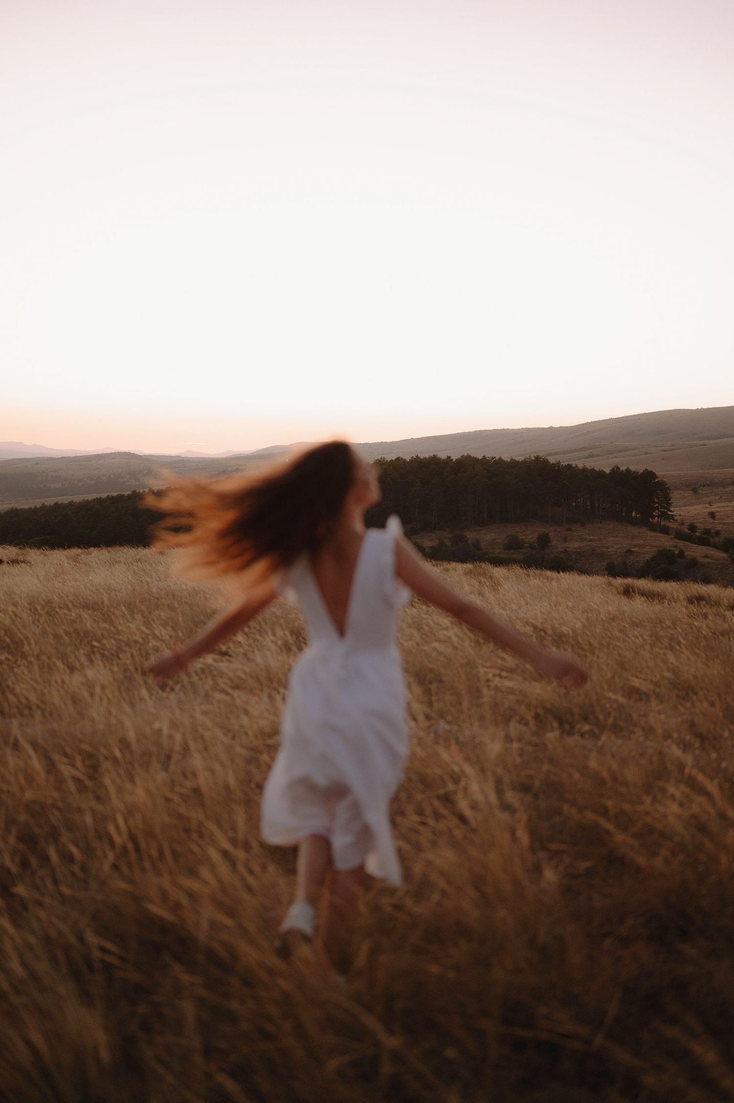 Someone in a white dress twirls freely through tall grass against a mountainous landscape at dusk.