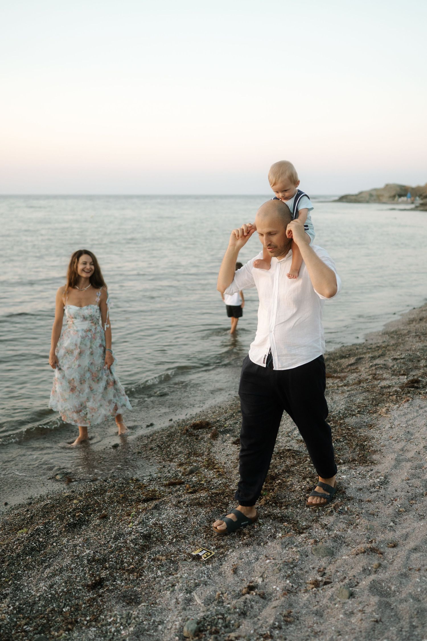 A couple playfully walks and dances together on a rocky beach at sunset with waves in the background.
