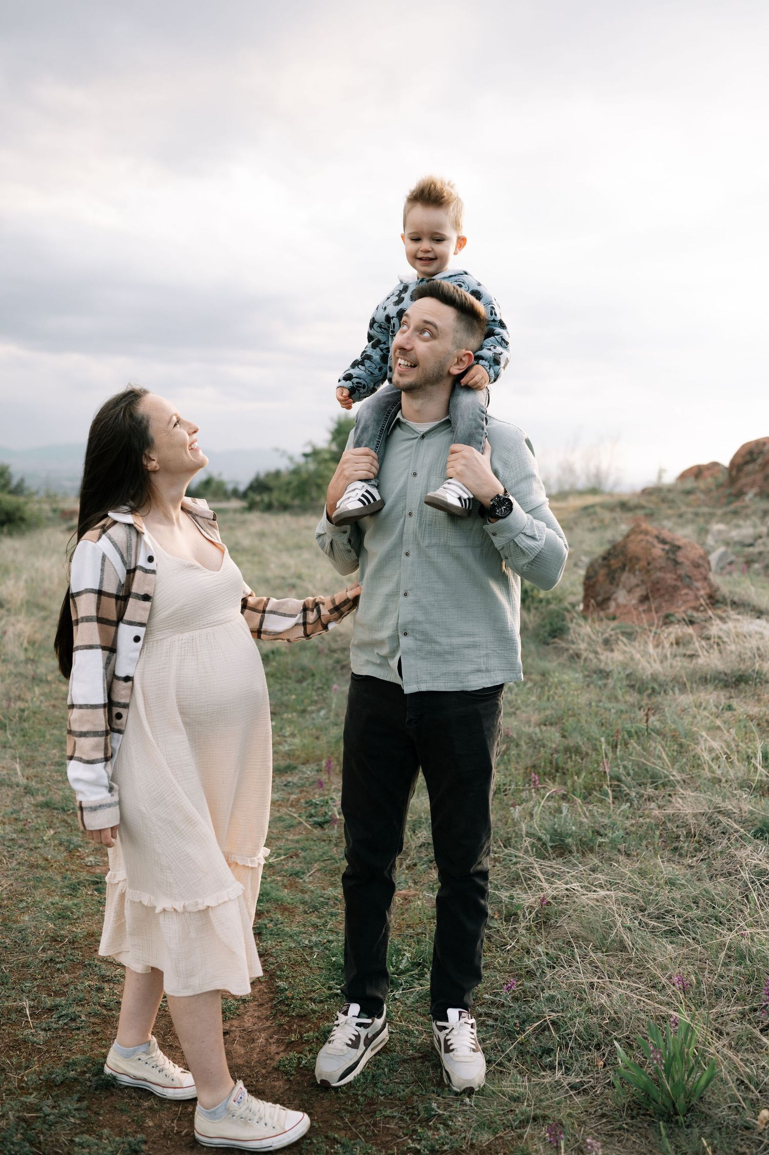 A family shares a joyful moment outdoors on a grassy hillside, with a pregnant woman in a dress and a child riding on shoulders.