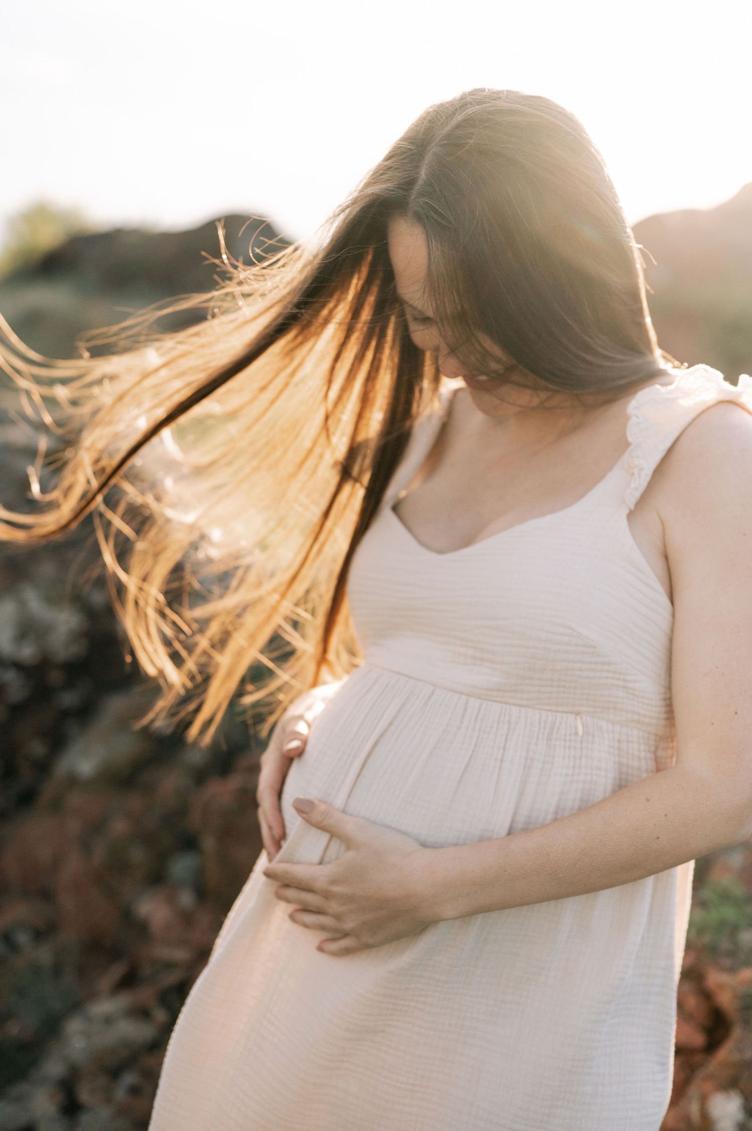 Soft backlit maternity portrait showing a flowing white dress and gentle embrace of baby bump at sunset.