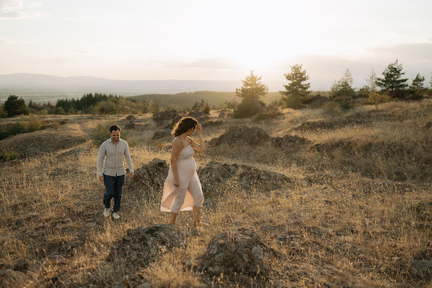 Two people stroll through dry grass on a hilltop during golden hour with mountains in the background.