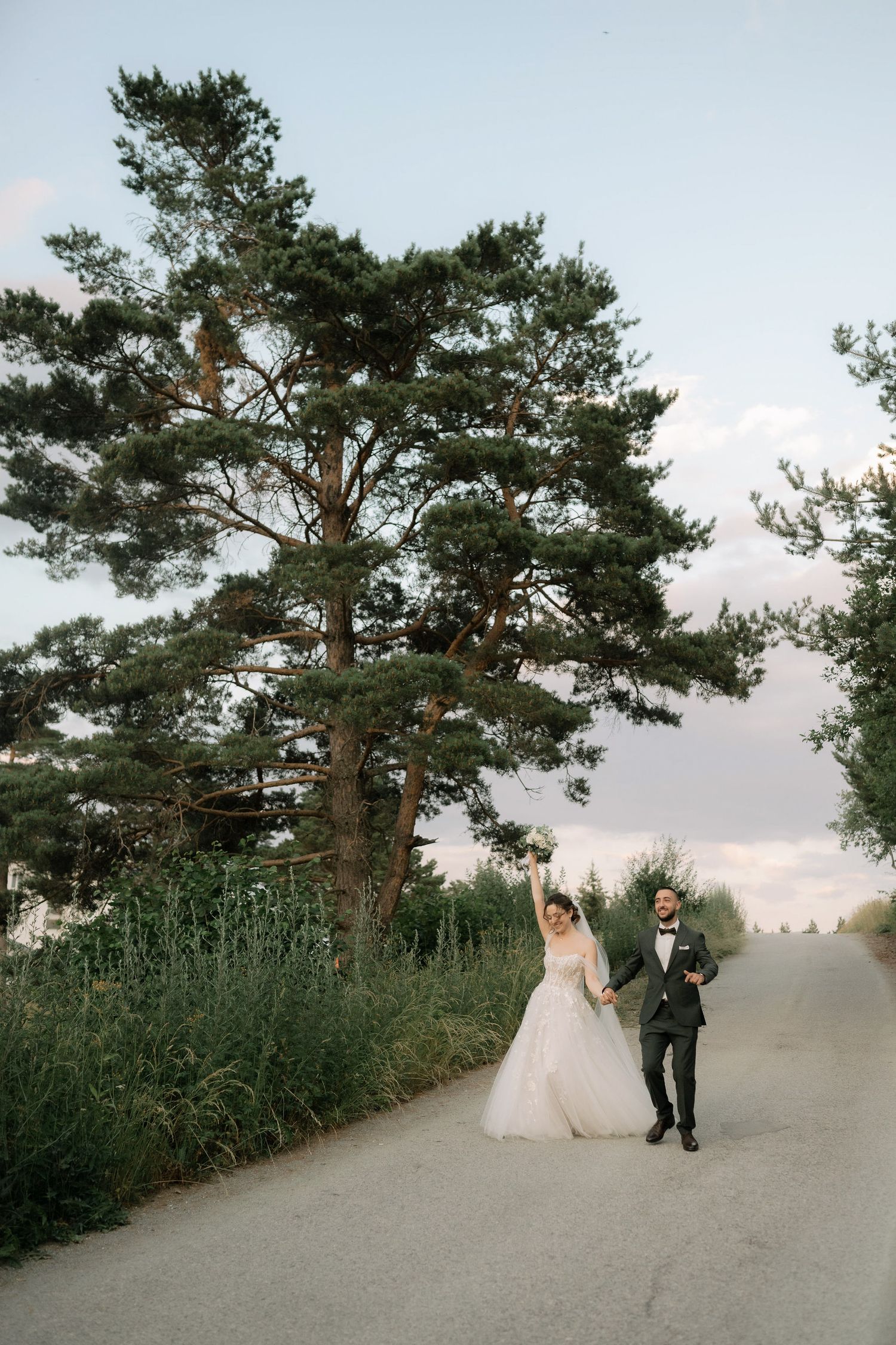 A couple walks along a scenic pine tree-lined path during a wedding photo session at sunset.
