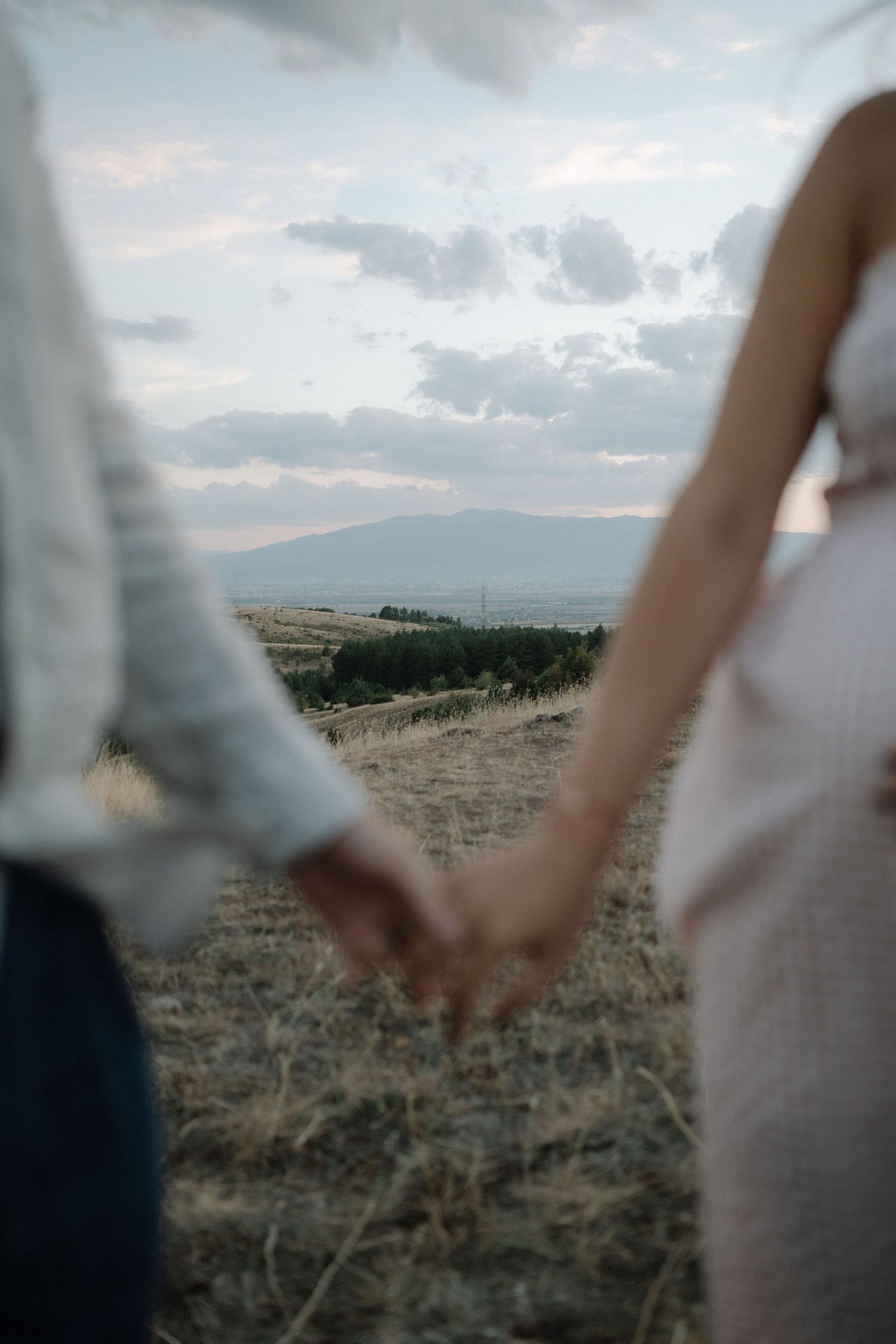 Couple holding hands while walking through a misty field at dusk with mountains in the distance.