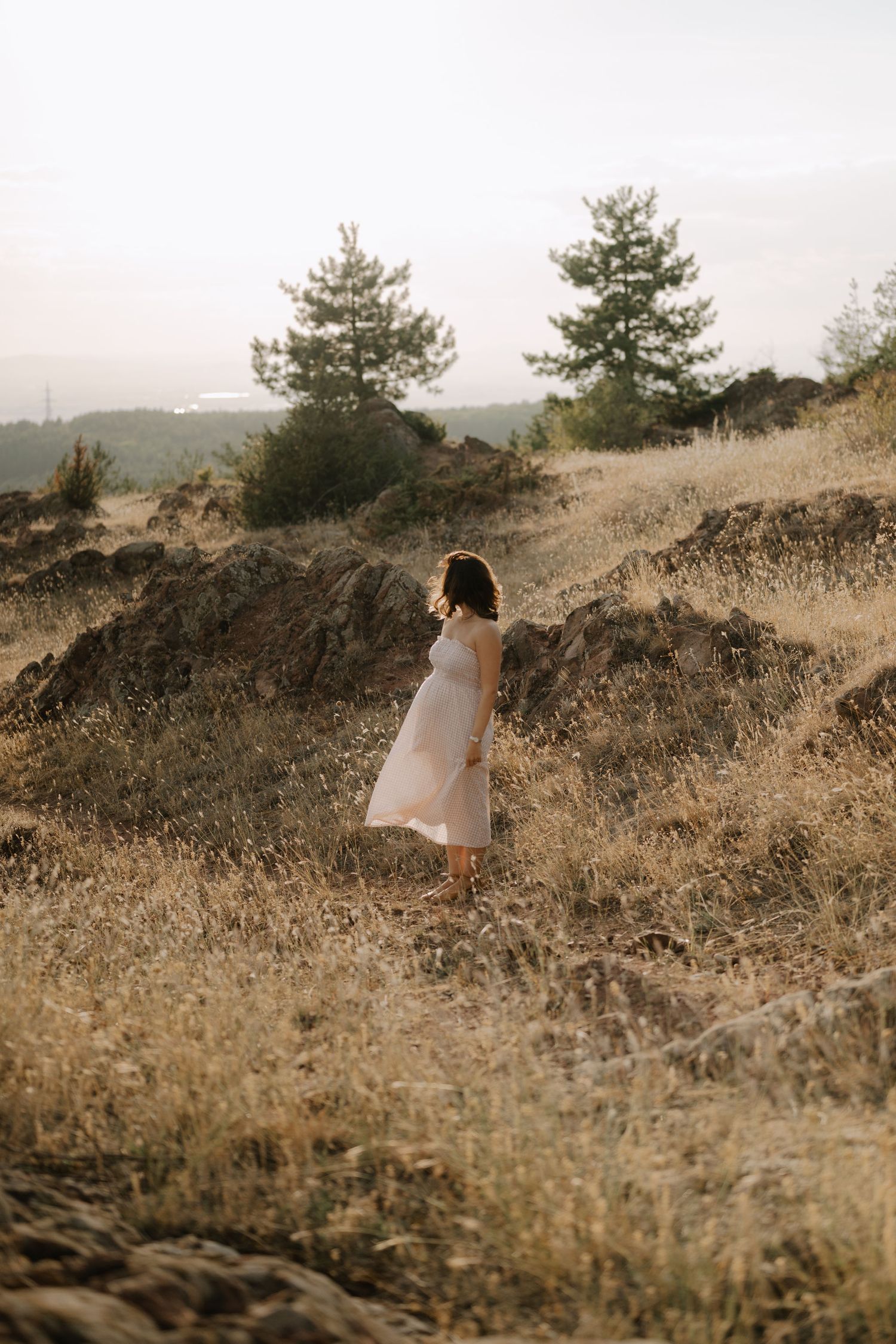 A figure in a white dress stands on a golden hillside with evergreen trees silhouetted against a misty mountain backdrop.