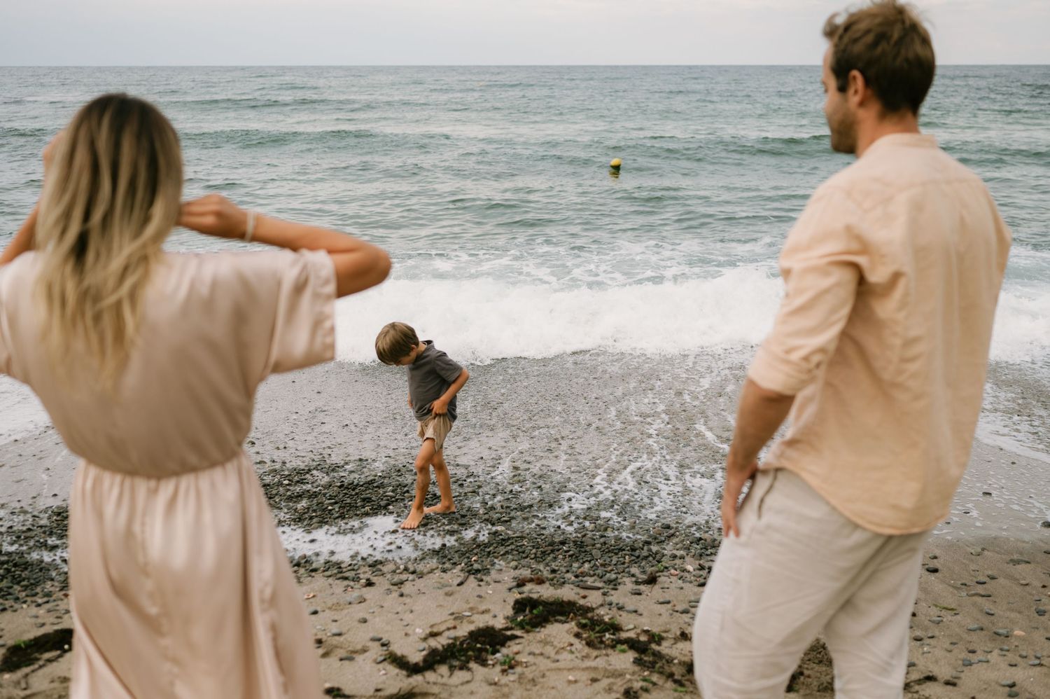 Family enjoying a beach day, parents watching child play in ocean waves on a cloudy day.