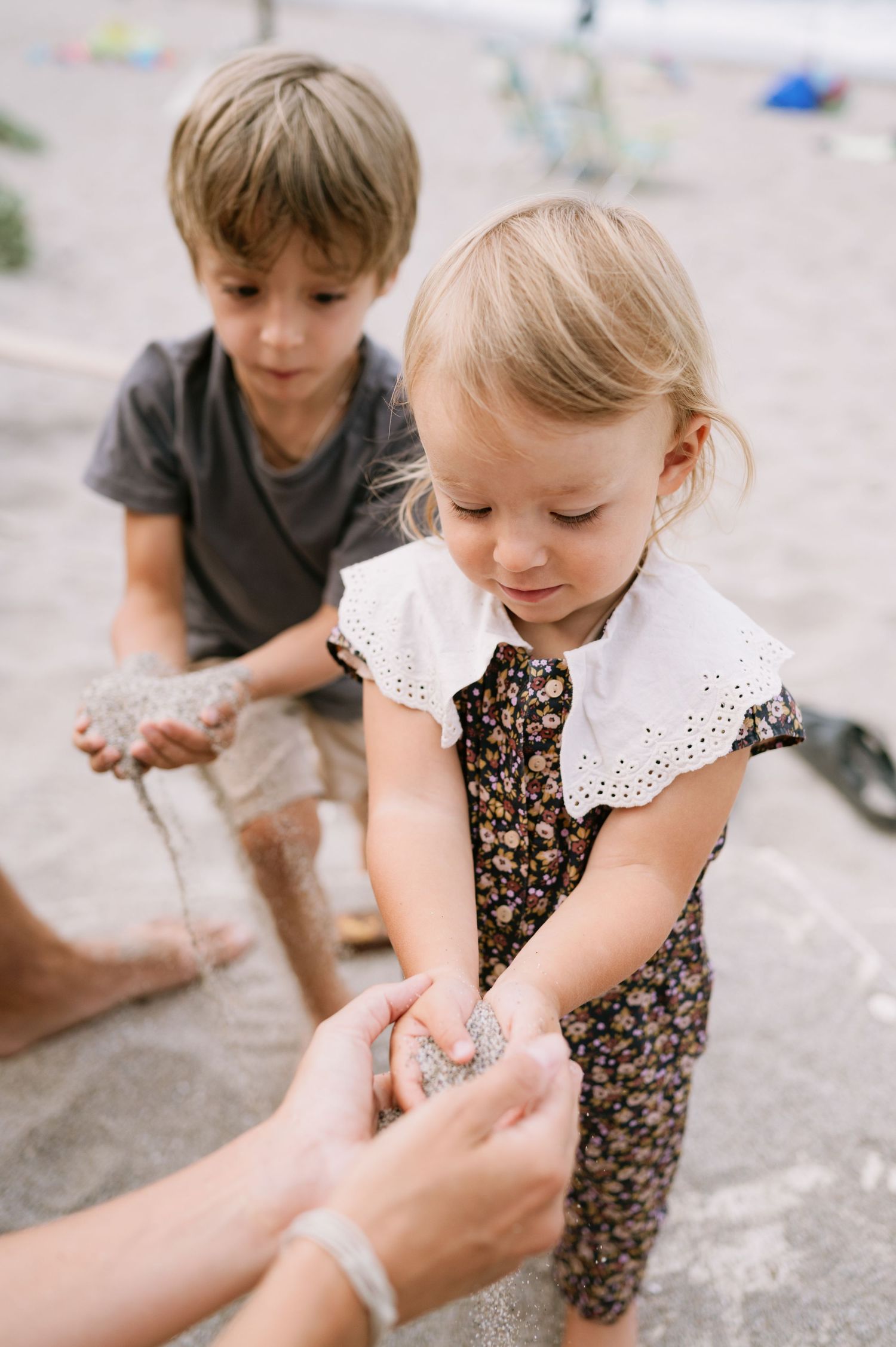 Two young children playing with sand on a beach, girl in floral romper reaching out to adult hands.