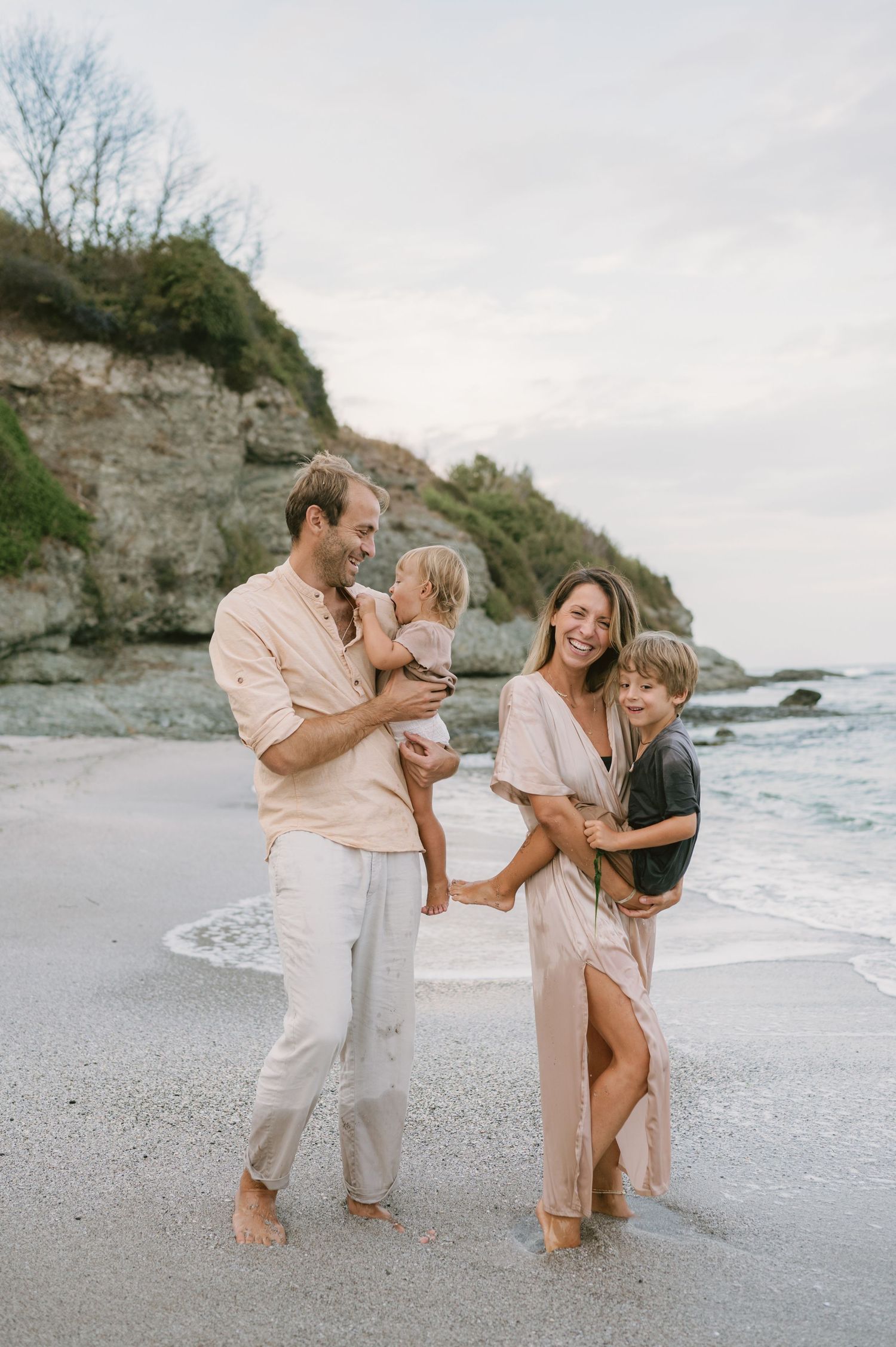 Happy family of four posing barefoot on a sandy beach with rocky cliffs, wearing neutral tones in a candid coastal portrait.
