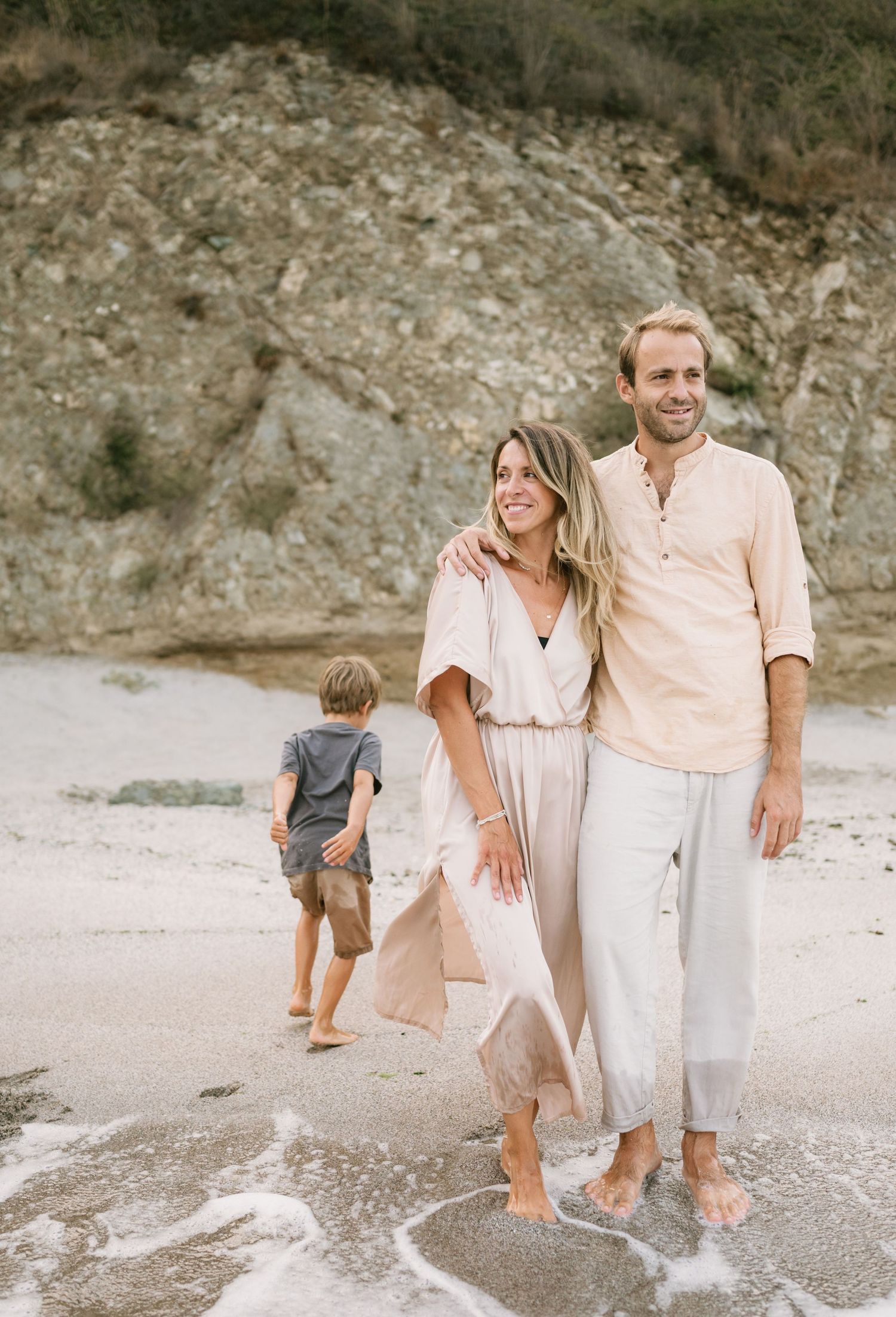 Couple with child on sandy beach, wearing neutral tones, smiling with rocky cliffs in background.