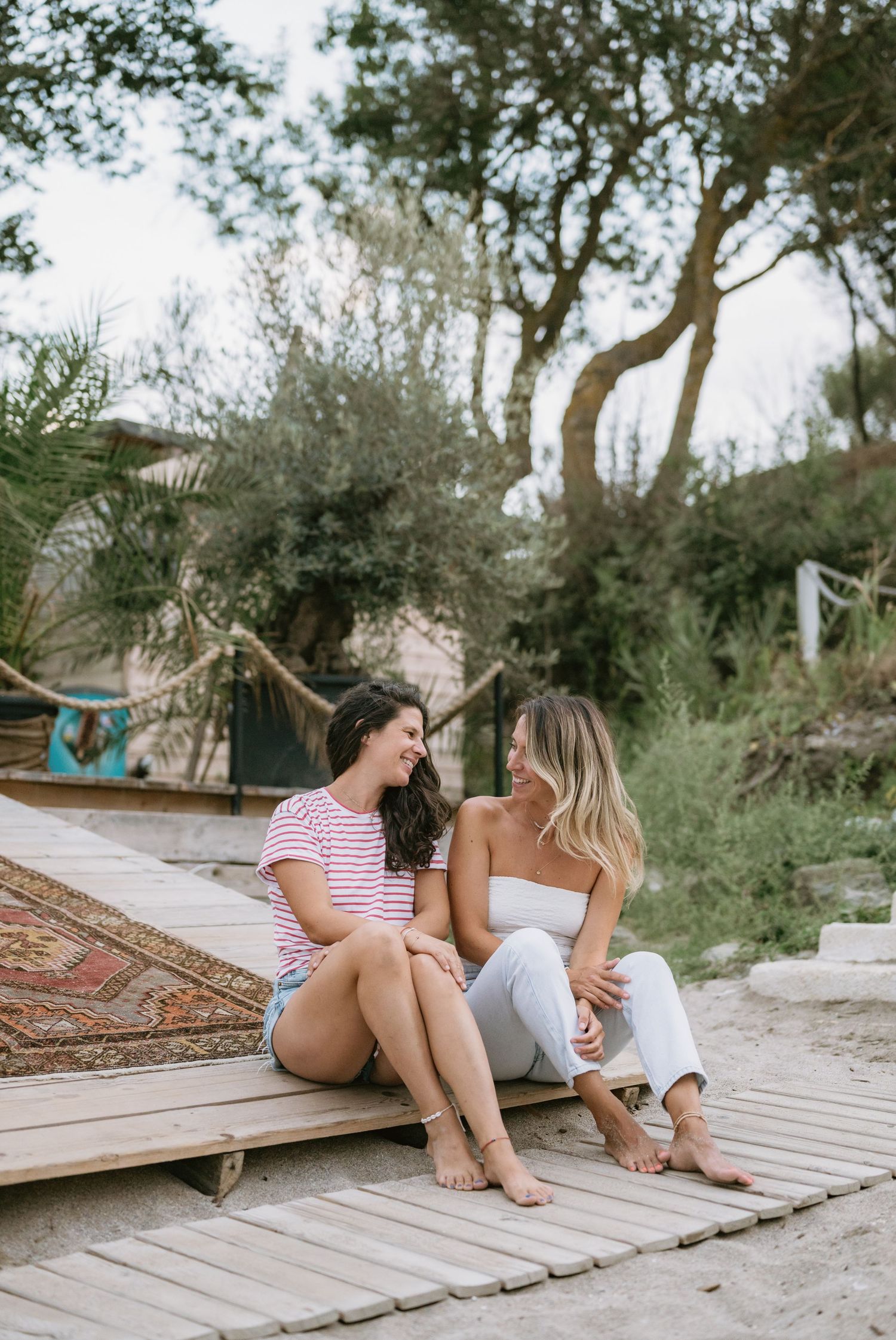 Two women sitting on a wooden deck, smiling at each other, surrounded by lush trees and a cozy outdoor setting.
