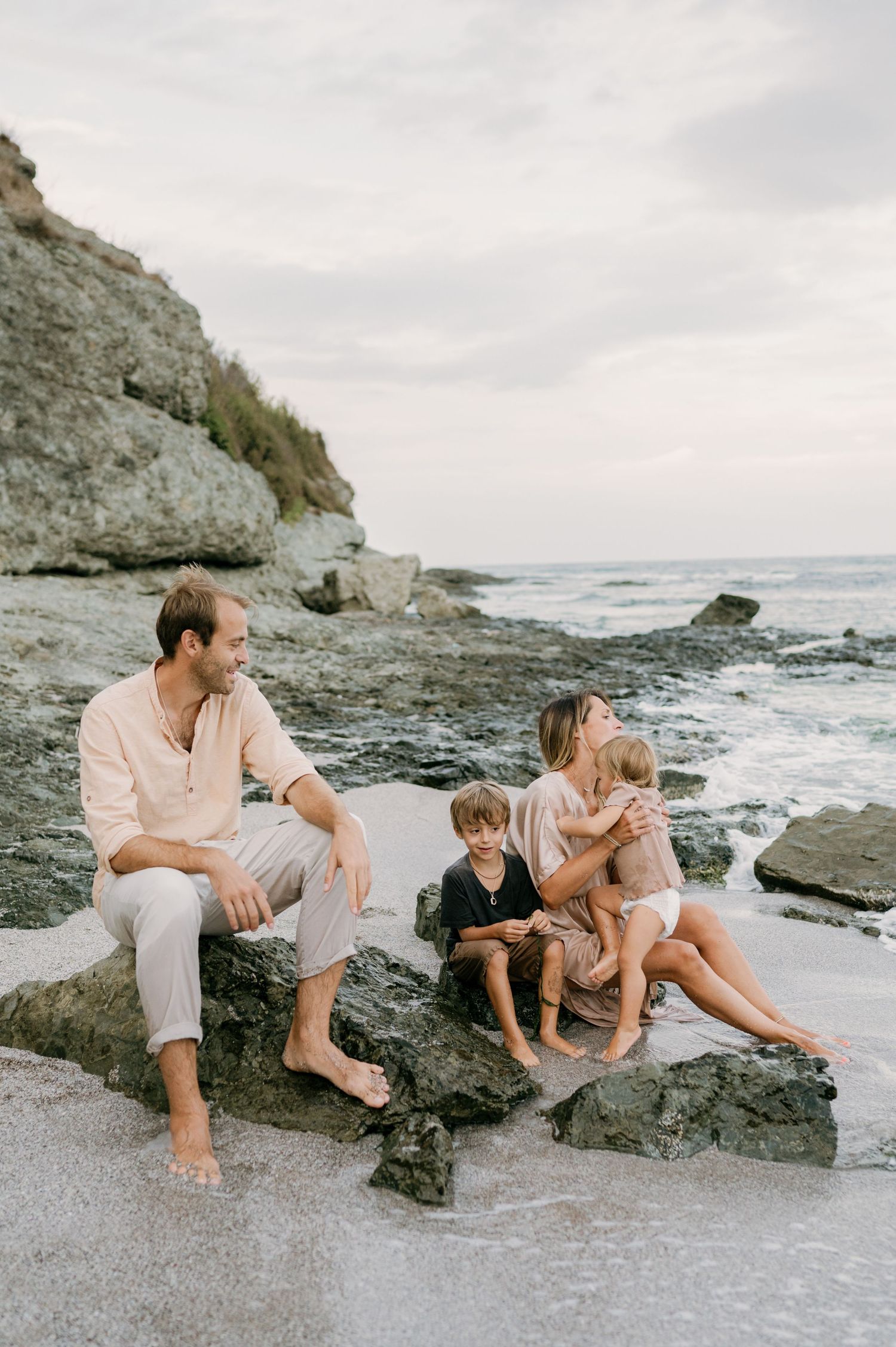 Family of four relaxing on rocky beach shoreline, parents and two children enjoying ocean waves at sunset.