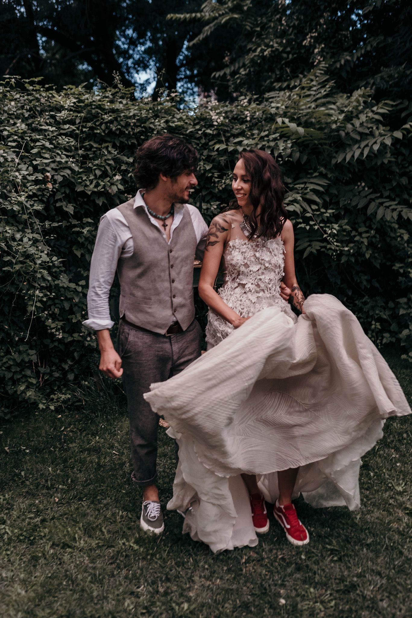 A couple in wedding attire playfully spins and dances together outdoors surrounded by lush greenery.