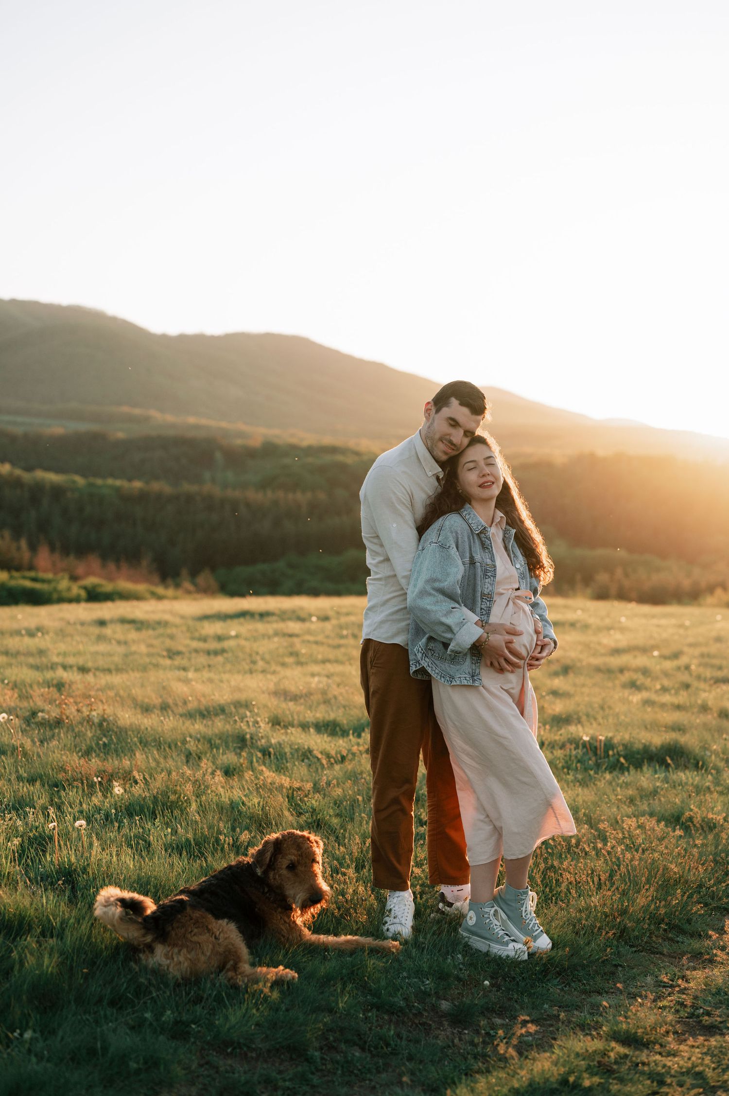 A couple embraces in a golden sunlit meadow with rolling hills and mountains in the background while their dog joins them.