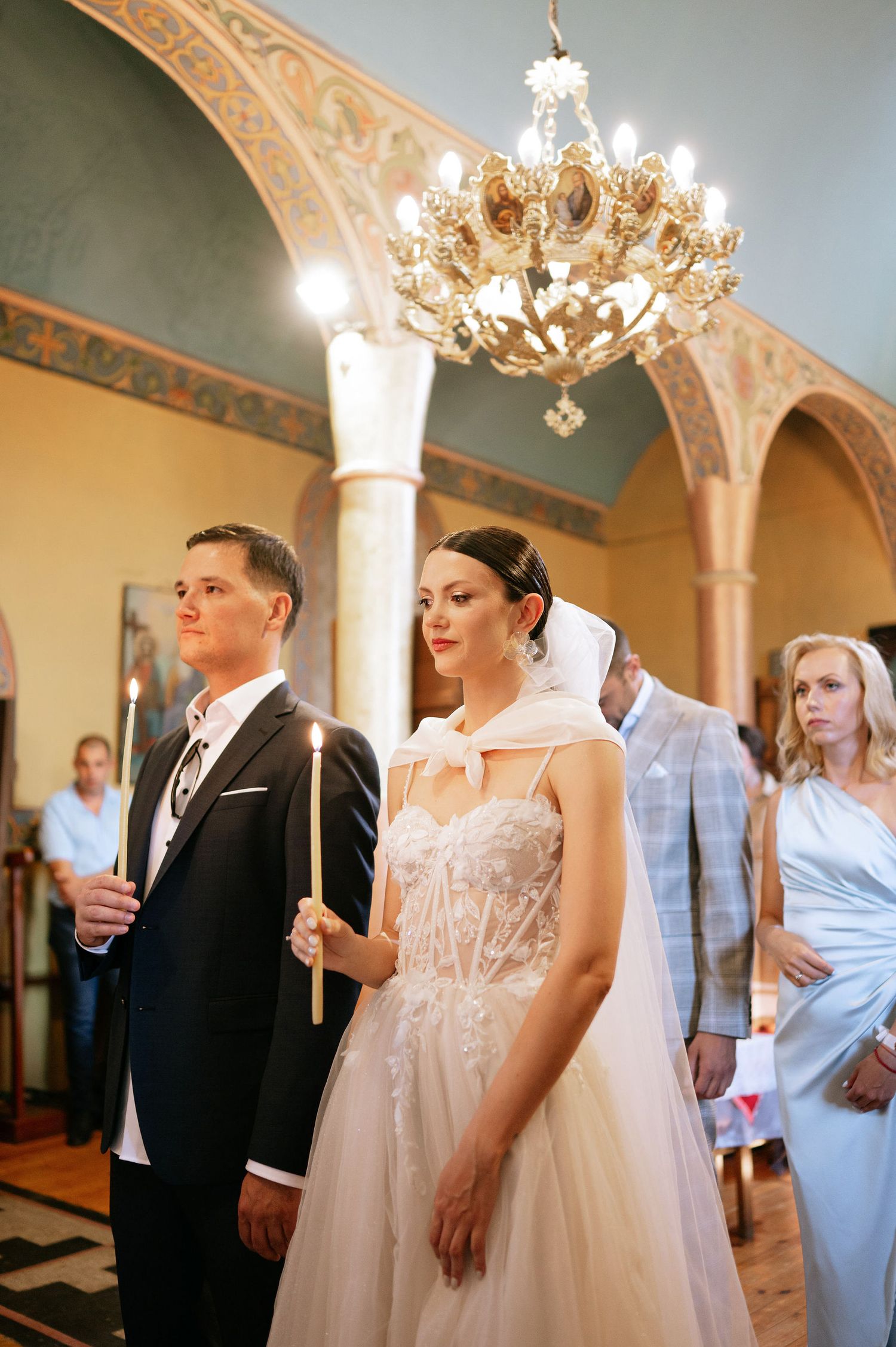 Wedding ceremony participants holding candles in an ornate church interior with chandeliers overhead.