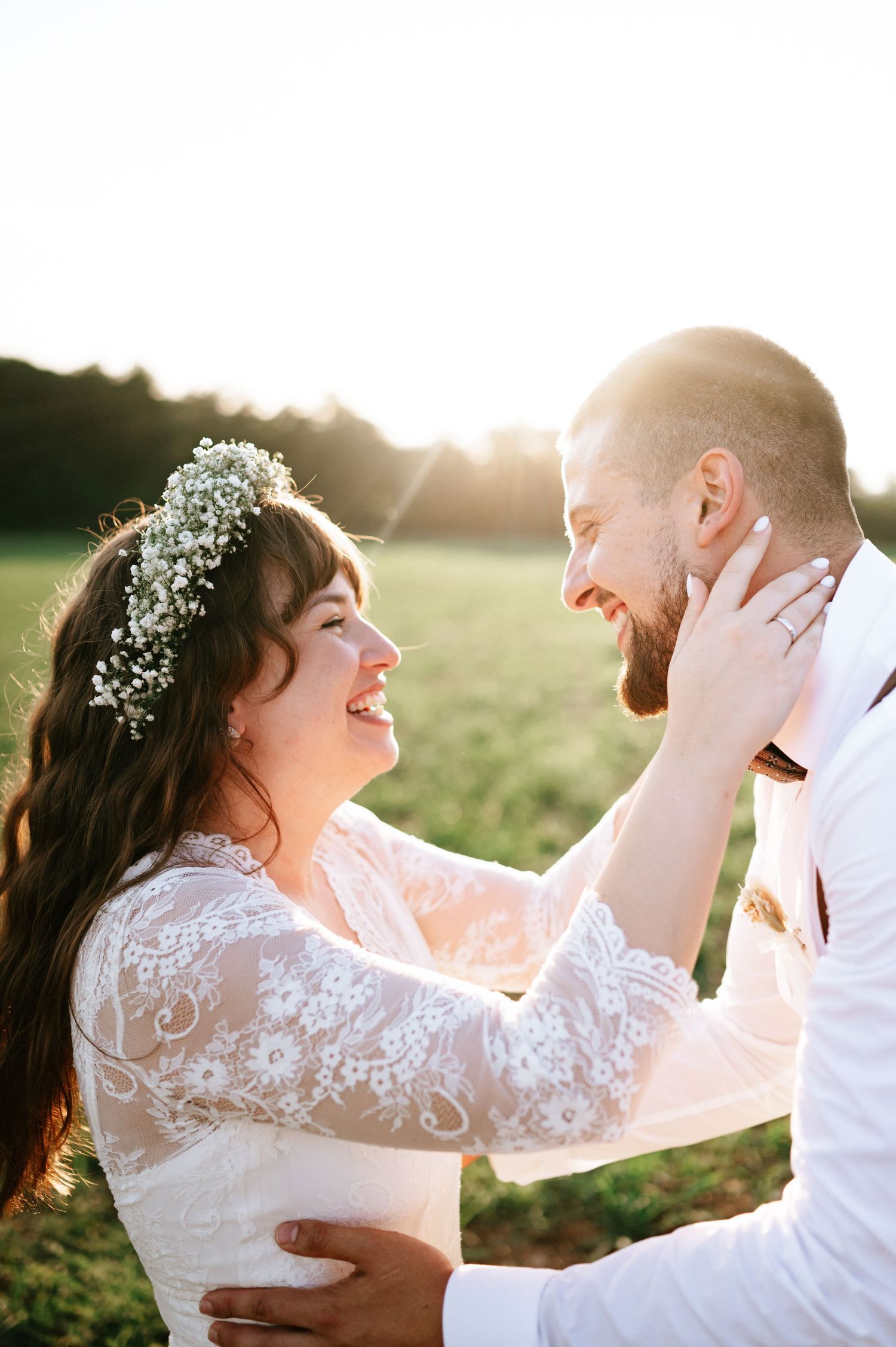 Candid wedding moment shows couple laughing together in sunlit field with baby's breath crown.