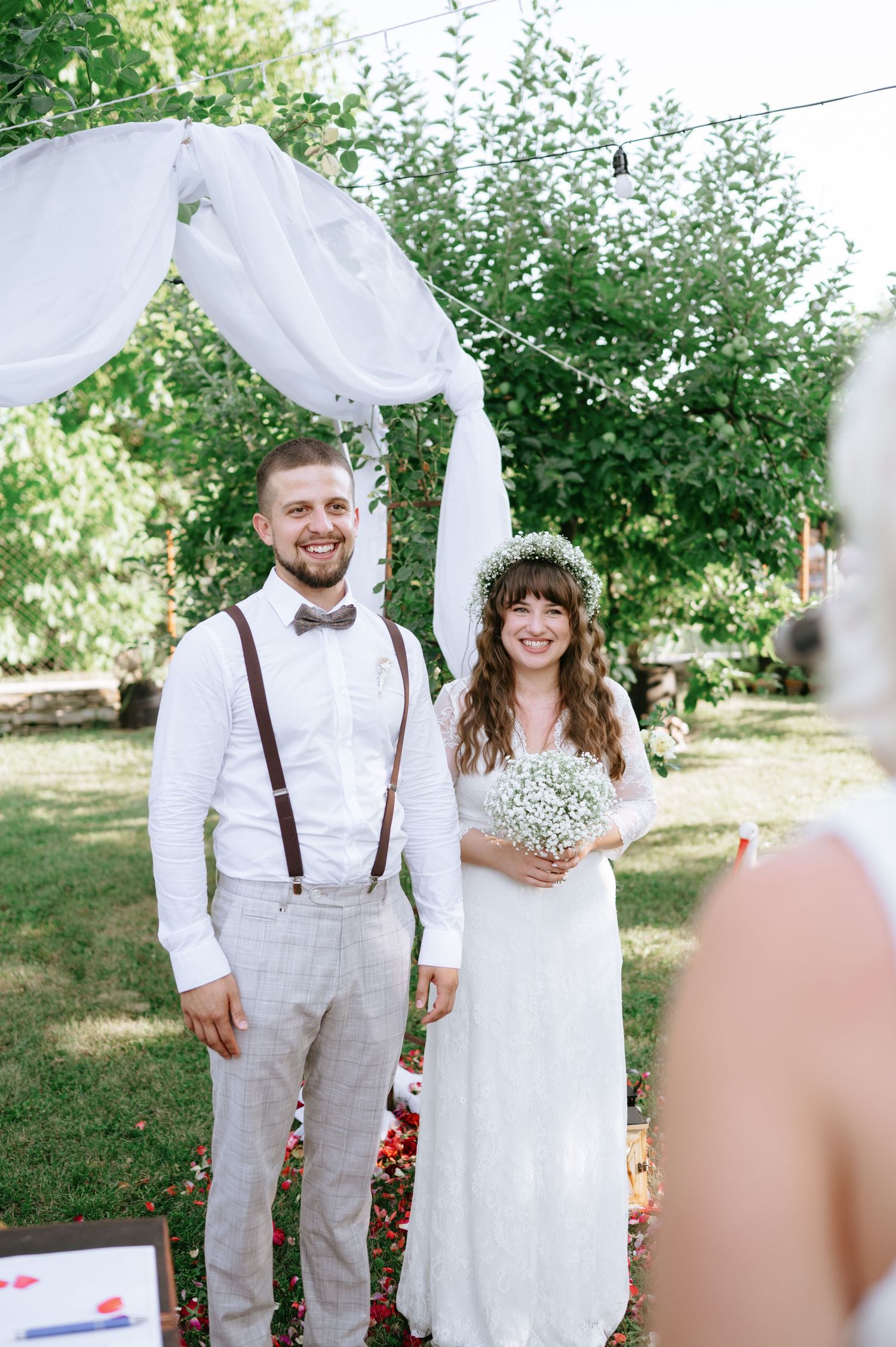 A couple shares a joyful moment under a white draped wedding arch surrounded by greenery.