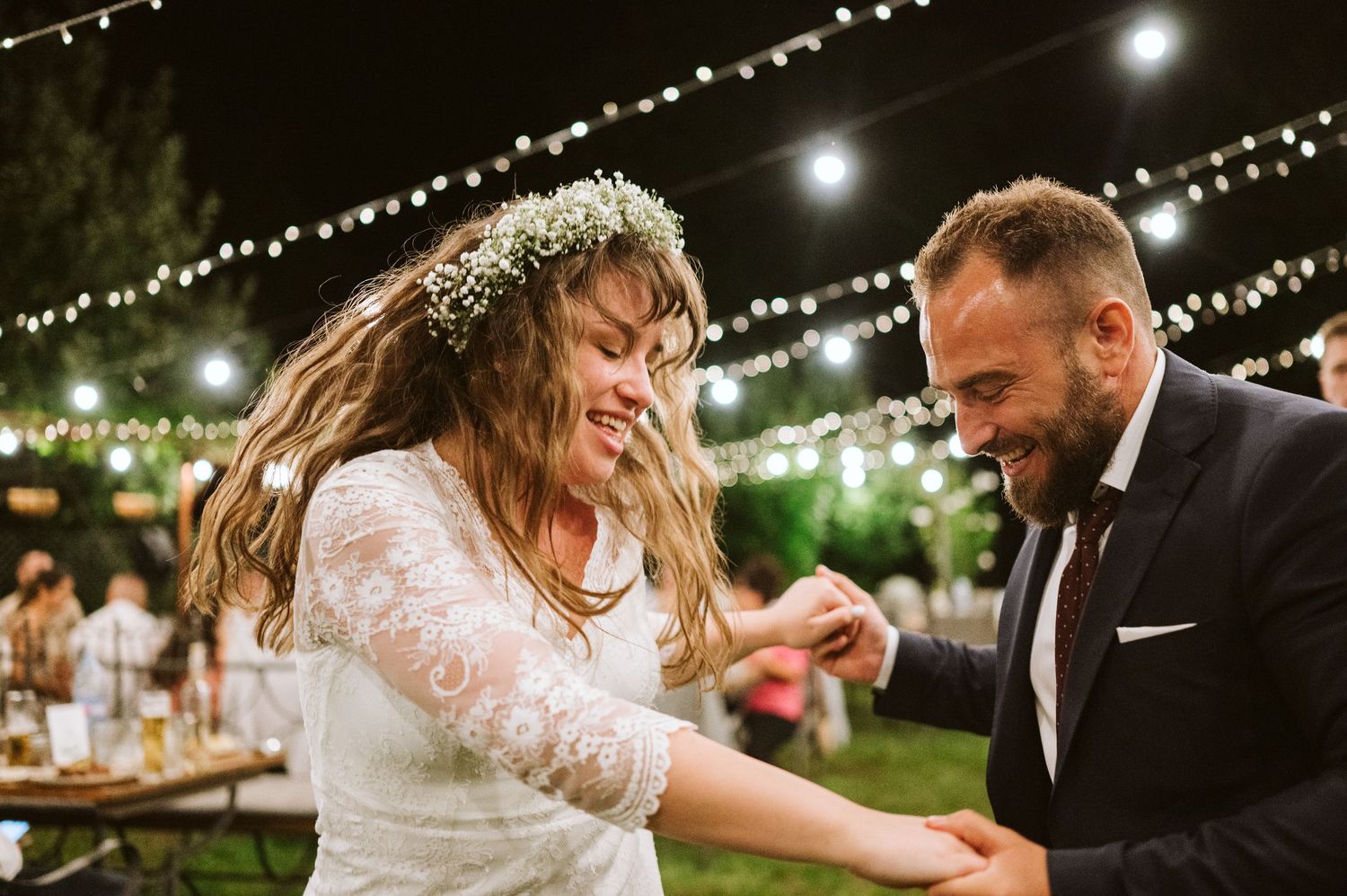 Newlyweds share playful moments while dancing under glowing string lights at outdoor reception.