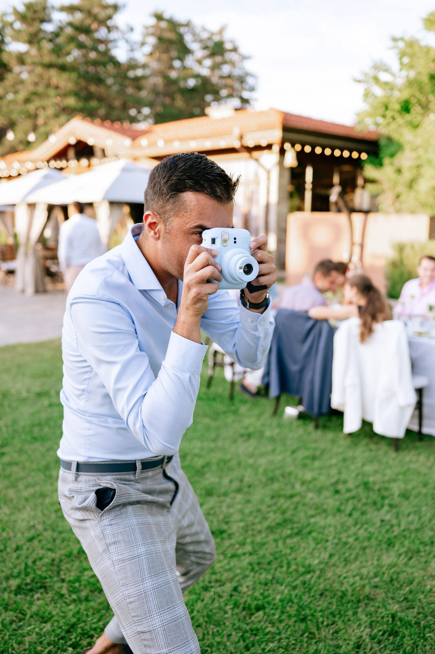 Wedding photographer in light blue shirt and gray pants taking photos at outdoor reception.