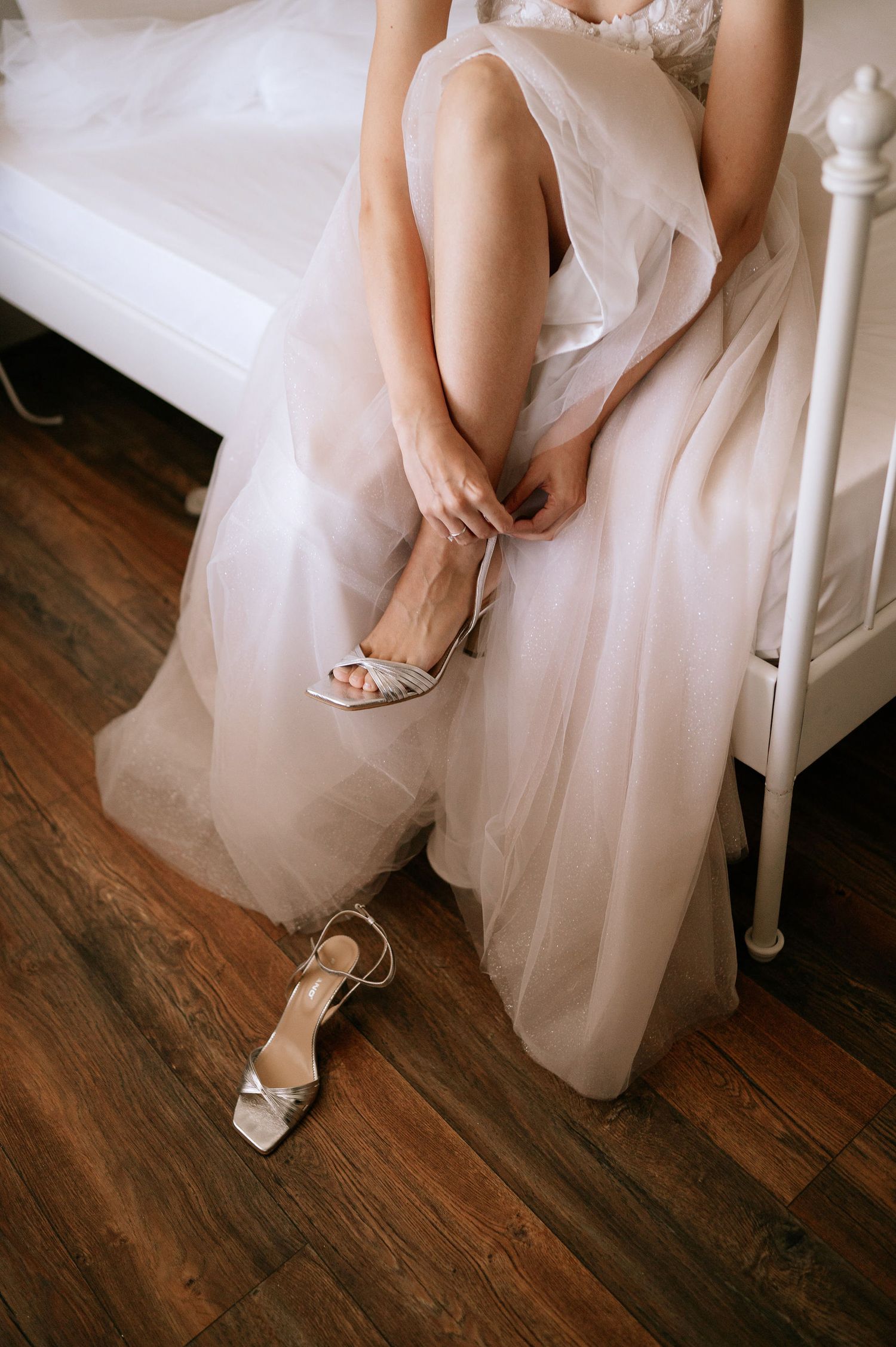 Close-up view of elegant bridal shoes and flowing white wedding dress on dark hardwood floors.
