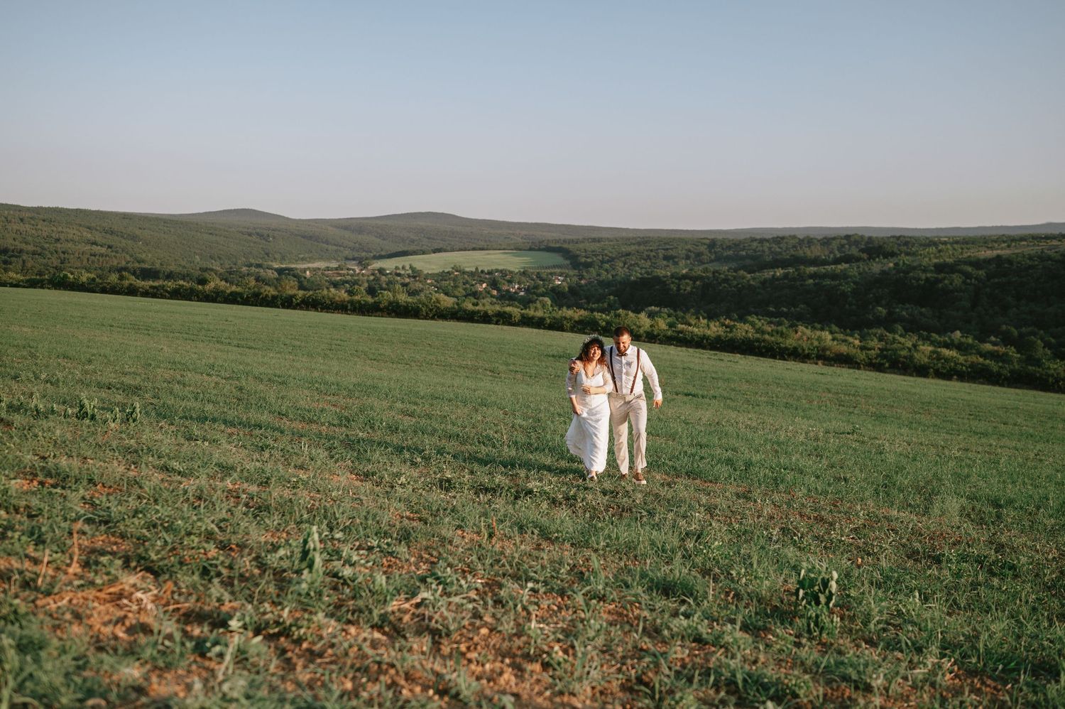 Two figures in white stand together on a vast green hillside with mountains in the distance.
