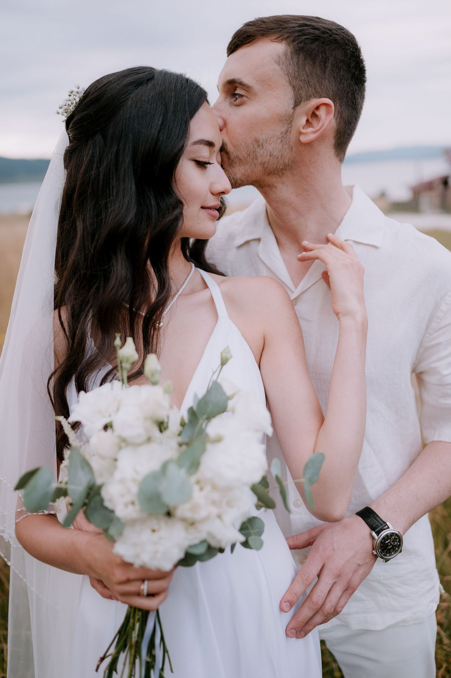 Newlyweds share tender moments while holding a white floral bouquet in nature.