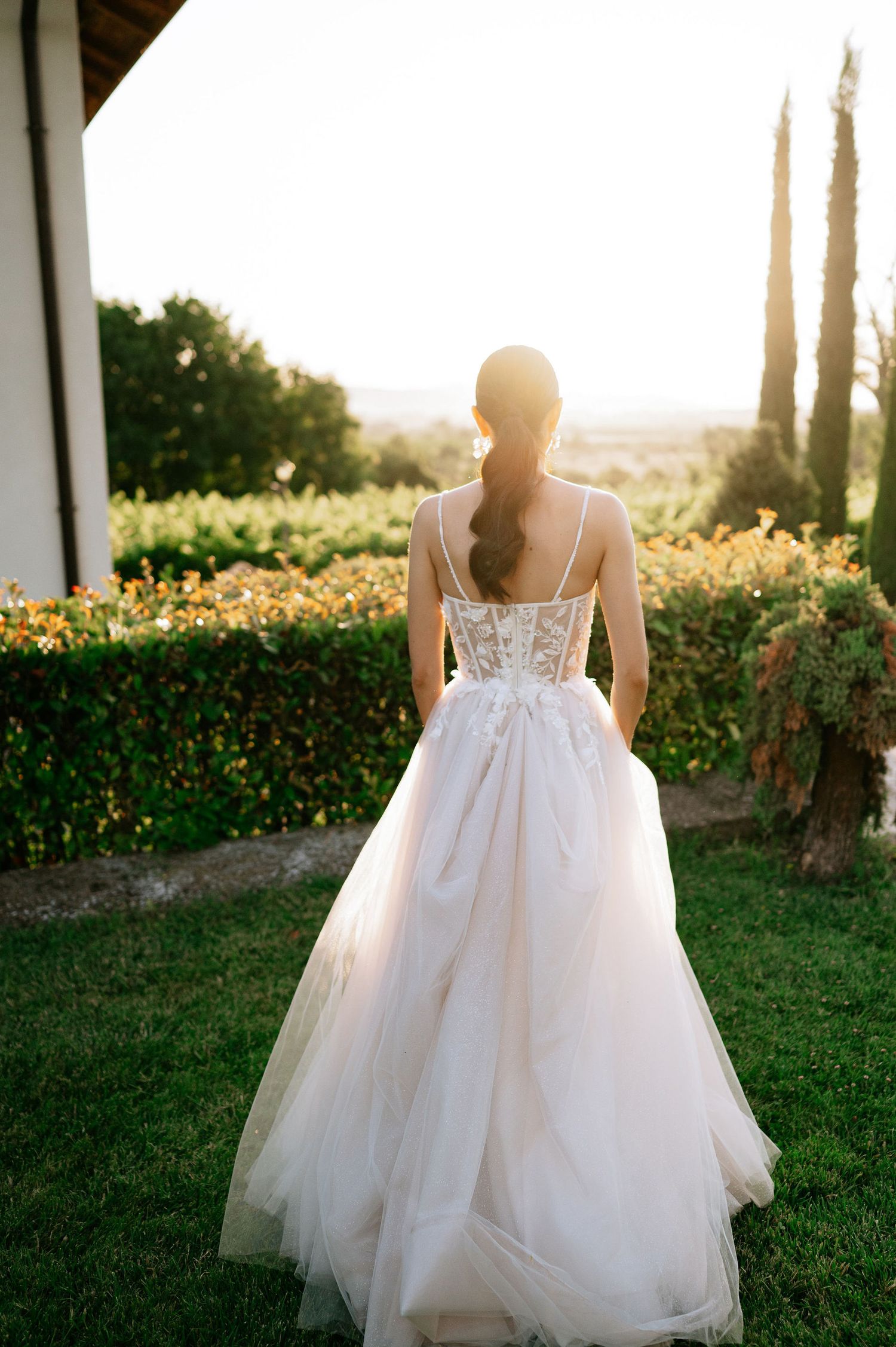 A flowing white wedding dress is showcased in golden sunlight among cypress trees and manicured gardens.